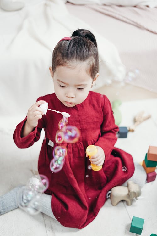 Girl in Red Long Sleeve Dress Sitting on White Floor Tiles Playing With Bubble Bubbles