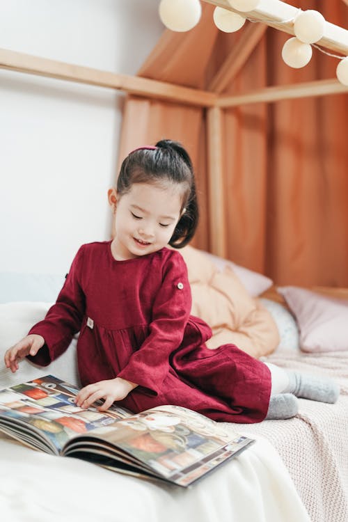 Free Girl in Red Dress Shirt Sitting on Bed Looking at Book Stock Photo