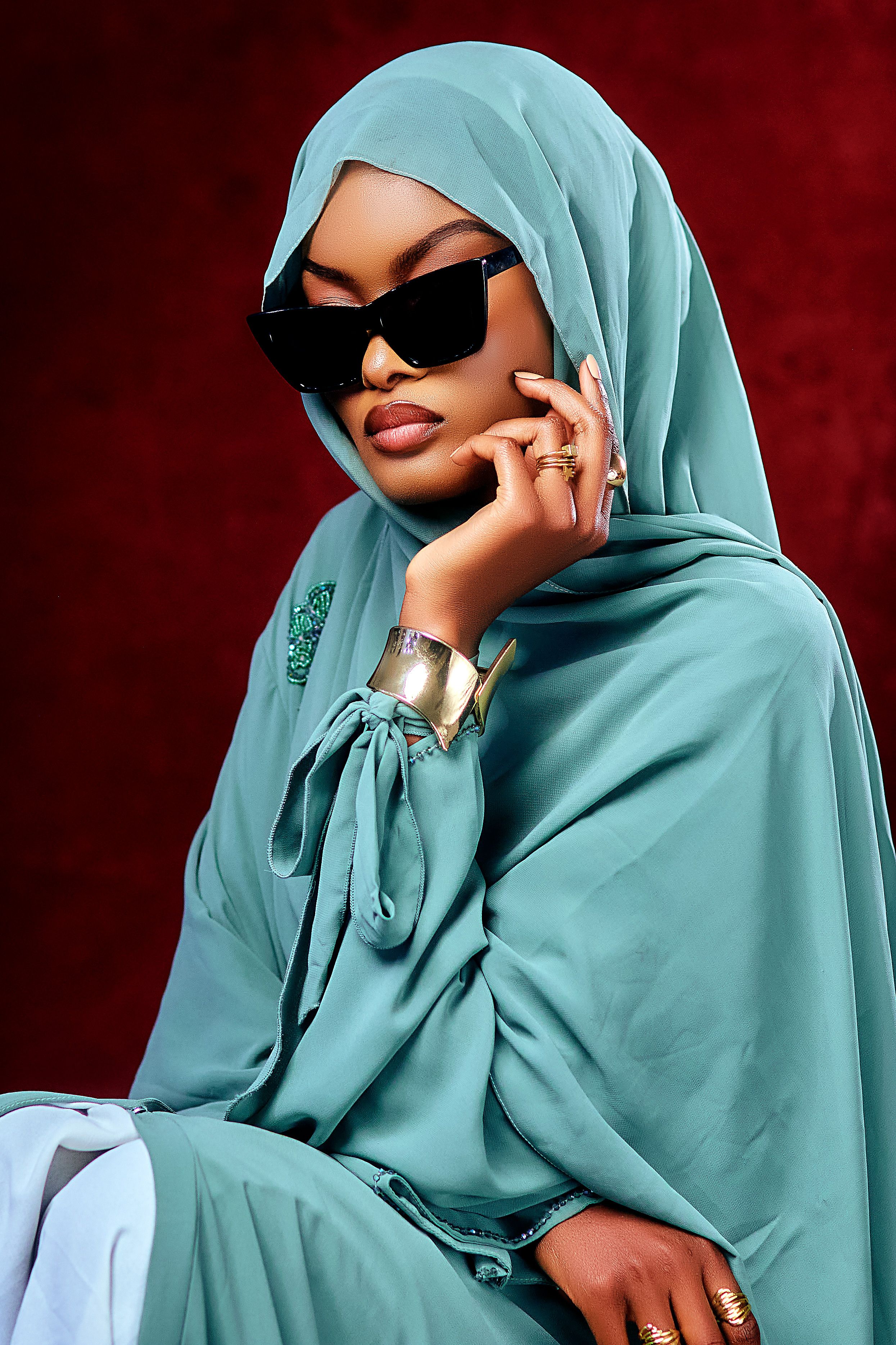 Stylish woman wearing a turquoise hijab and sunglasses against a rich red background.