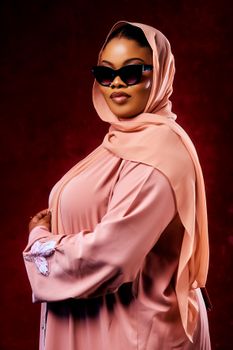 Stylish woman in a peach abaya and hijab with sunglasses, striking a pose against a dark red background.