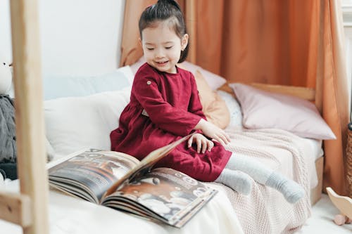 Girl in Red Long Sleeve Dress Sitting on Bed Reading Book 