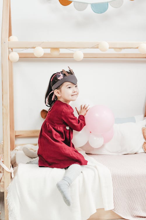 Girl in Red Long Sleeve Dress Sitting on Bed Playing with Balloons