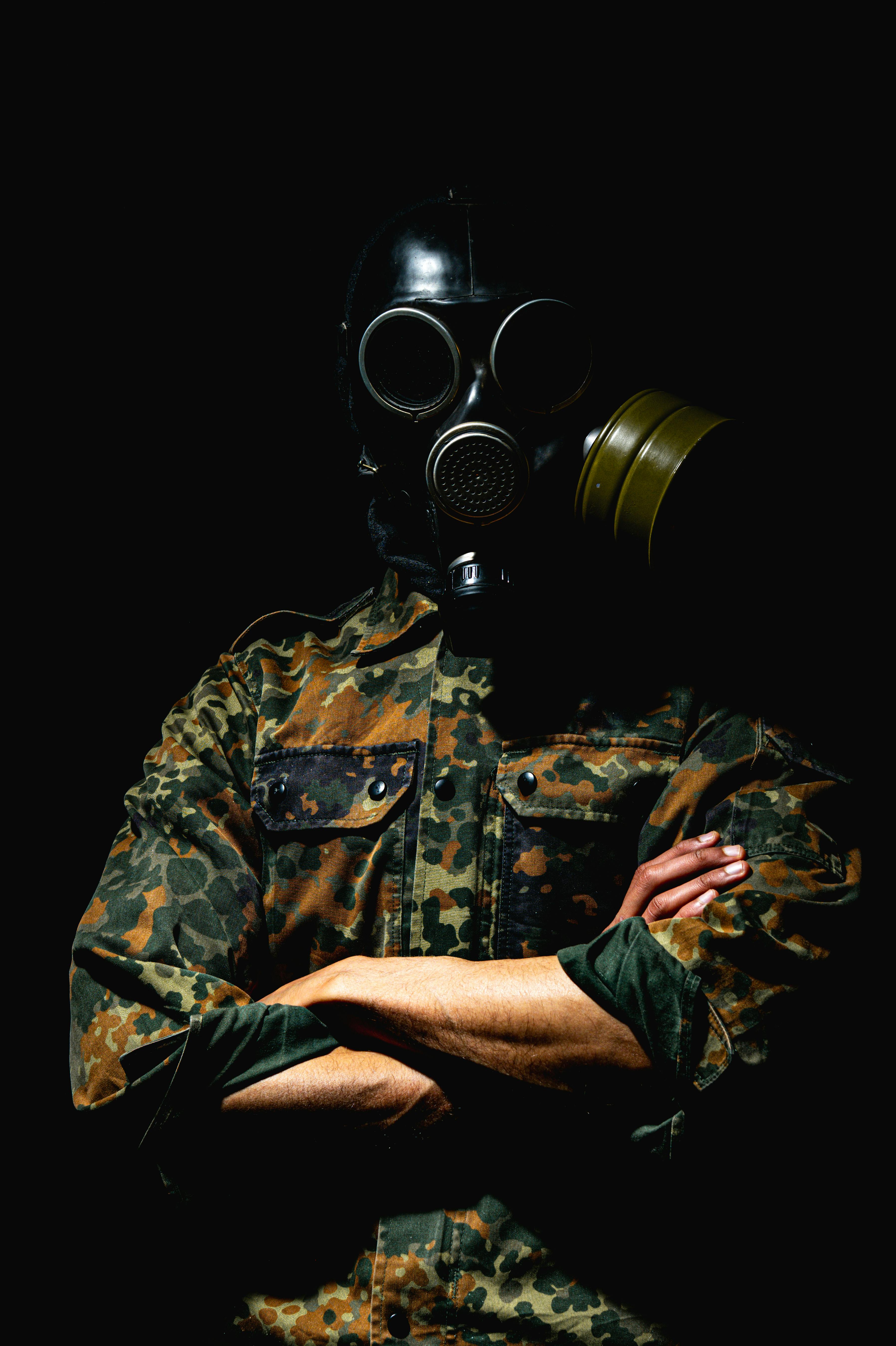 ingyenes Egy álcázóruhás és gázálarcban lévő személy keresztbe tett karokkal sötét háttér előtt. Stockfotó