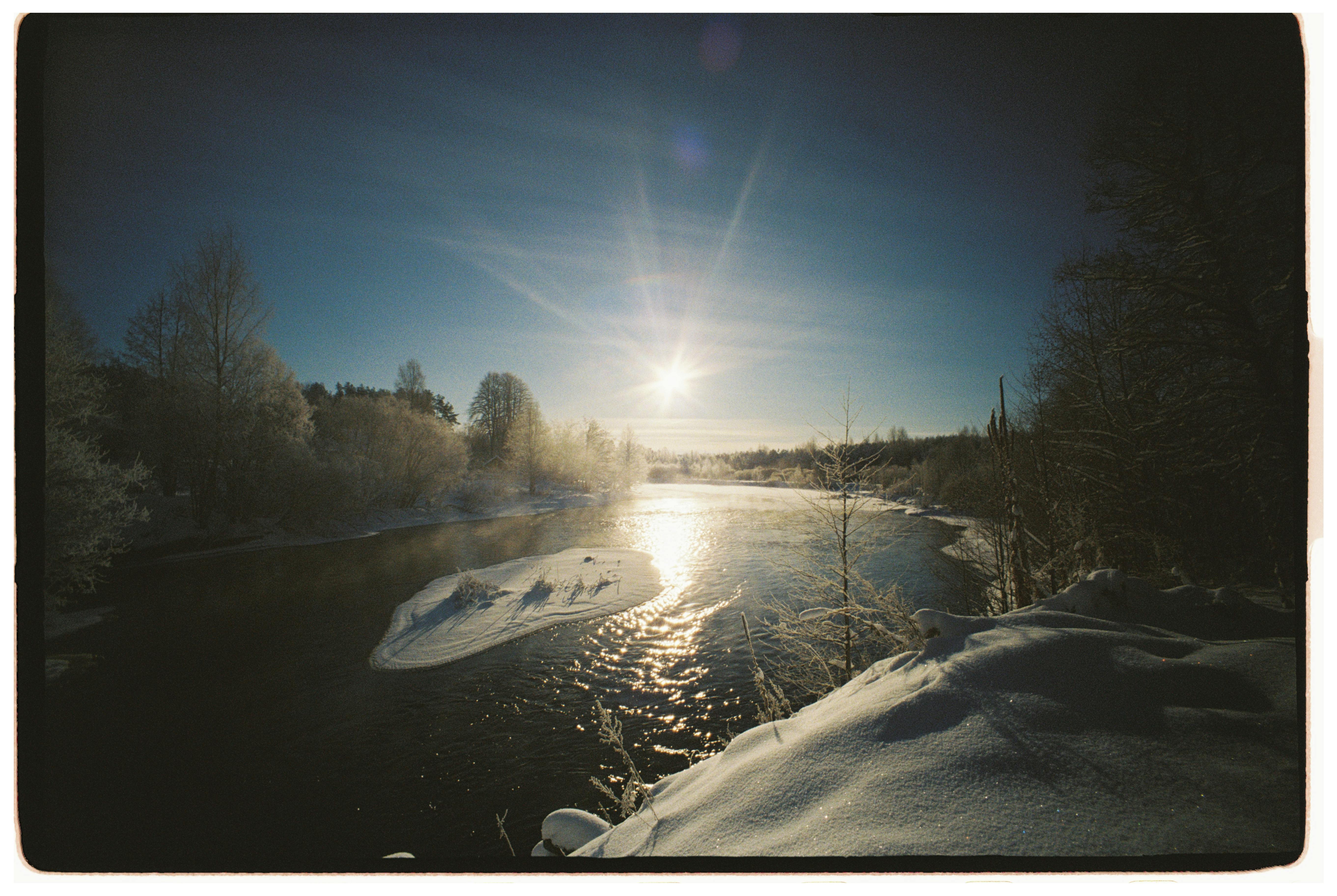 Безкоштовне Приголомшлива засніжена річкова сцена з яскравим зимовим сонцем, що відбивається від води. стокове фото