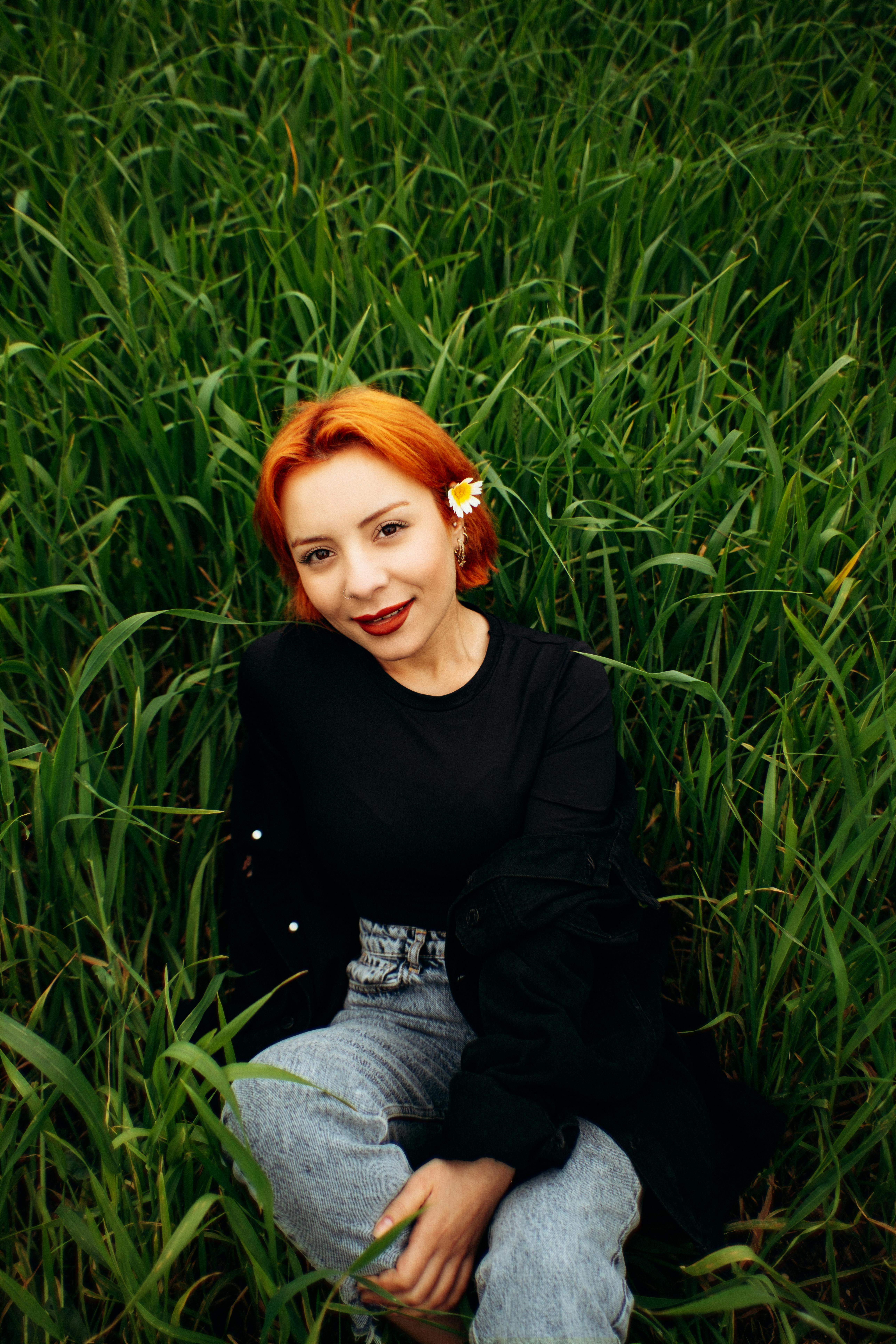 Безкоштовне Усміхнена жінка з рудим волоссям і квіткою в пишній траві, розслаблена на свіжому повітрі. стокове фото