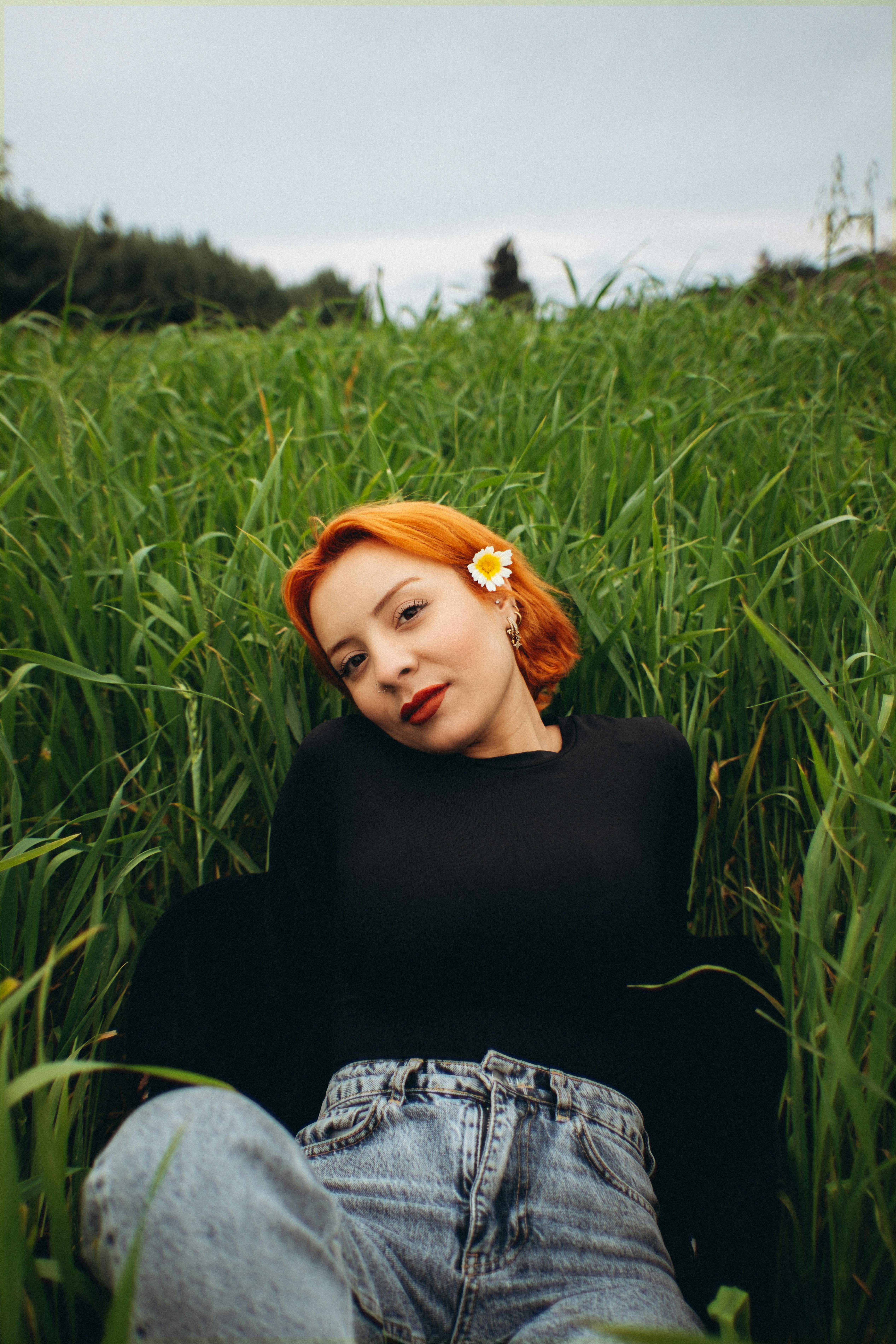 免費 有著橘色頭髮、手捧雛菊的年輕女子，在戶外高高的草叢中放鬆身心。 圖庫相片