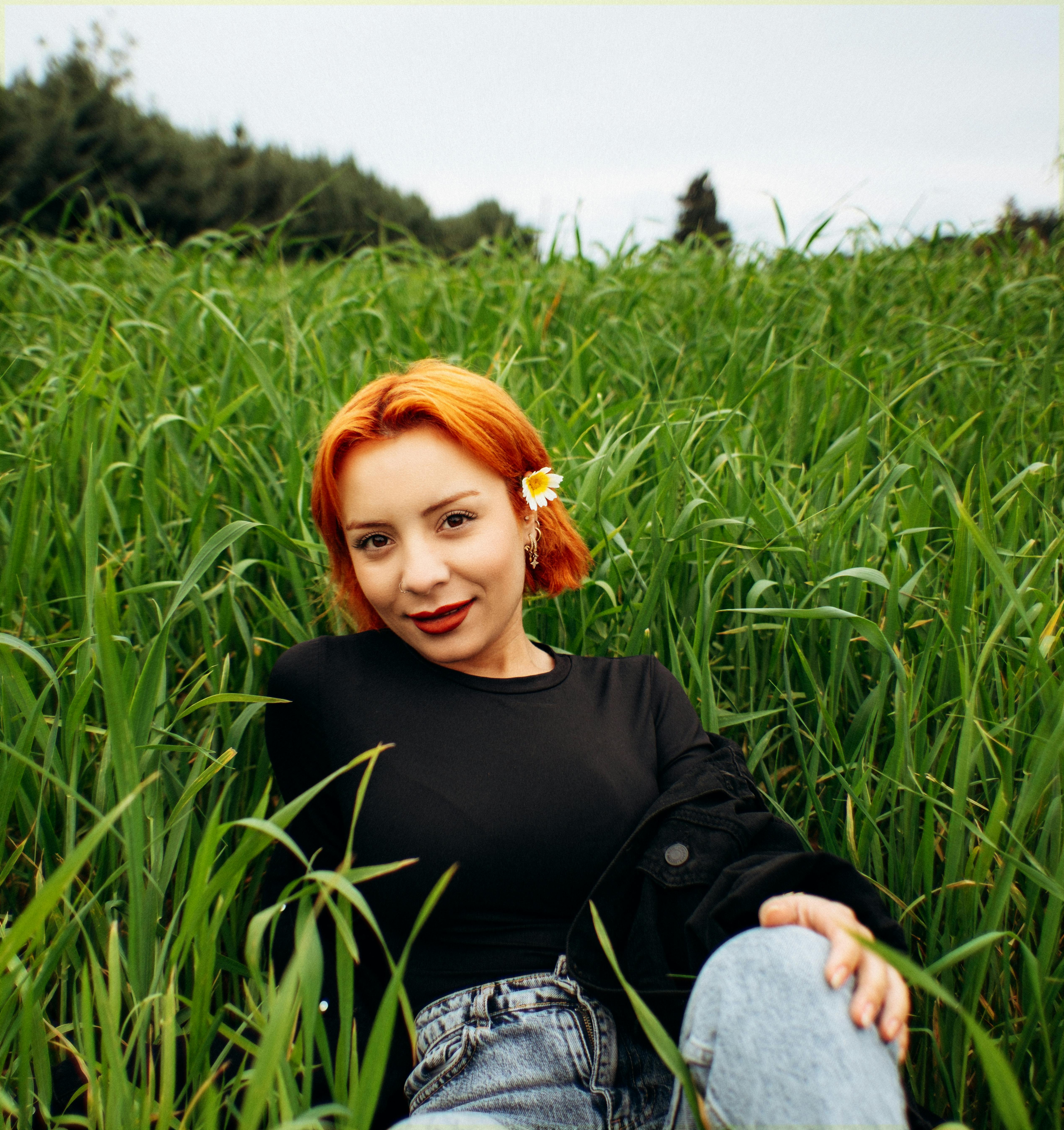 ฟรี หญิงสาวผมแดงยิ้มแย้มขณะนั่งอยู่ในทุ่งหญ้าเขียวชอุ่ม เพลิดเพลินกับธรรมชาติ คลังภาพถ่าย