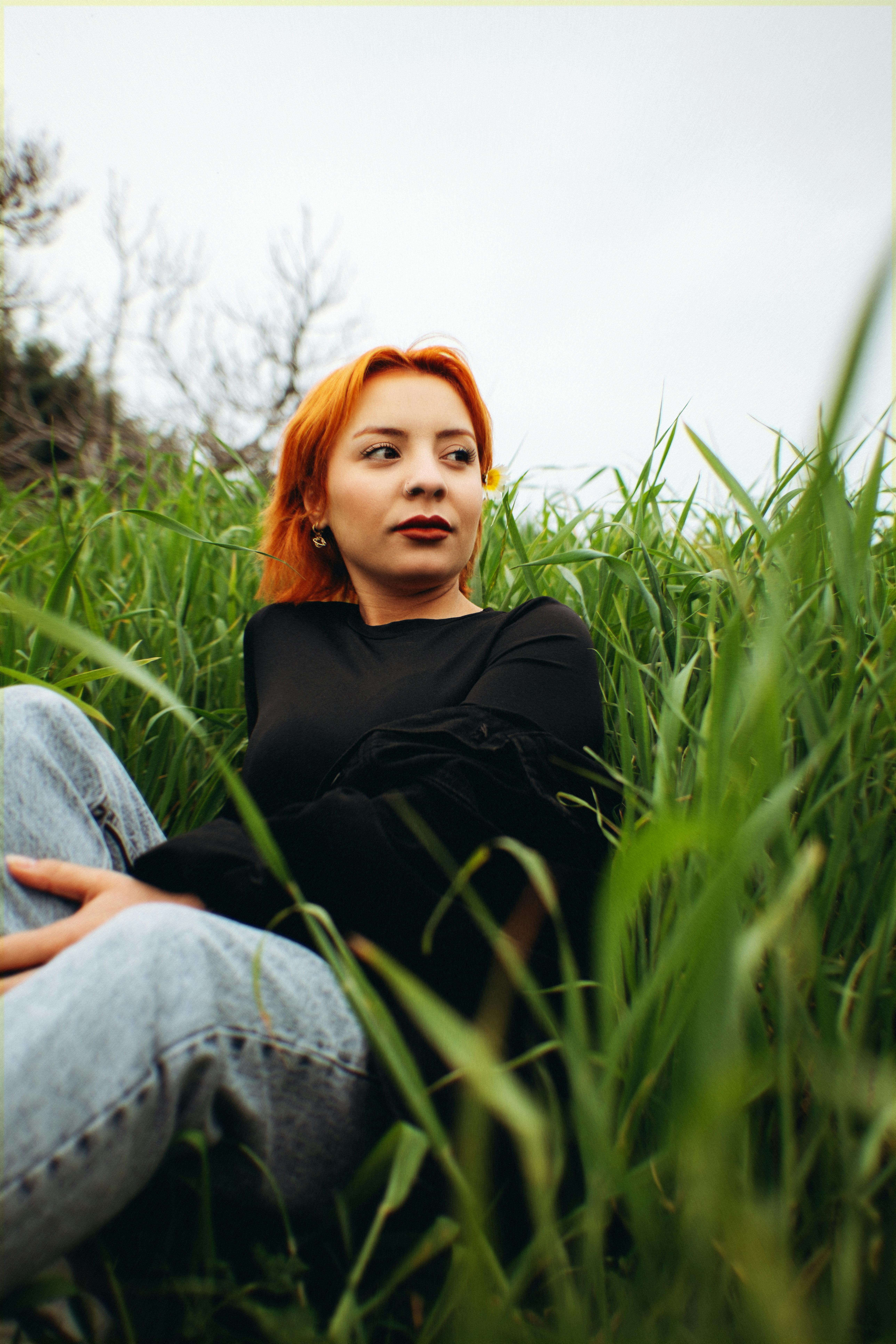 ingyenes Egy vörös hajú nő pihen egy mezőn, magas zöld fű veszi körül, felhős ég alatt. Stockfotó