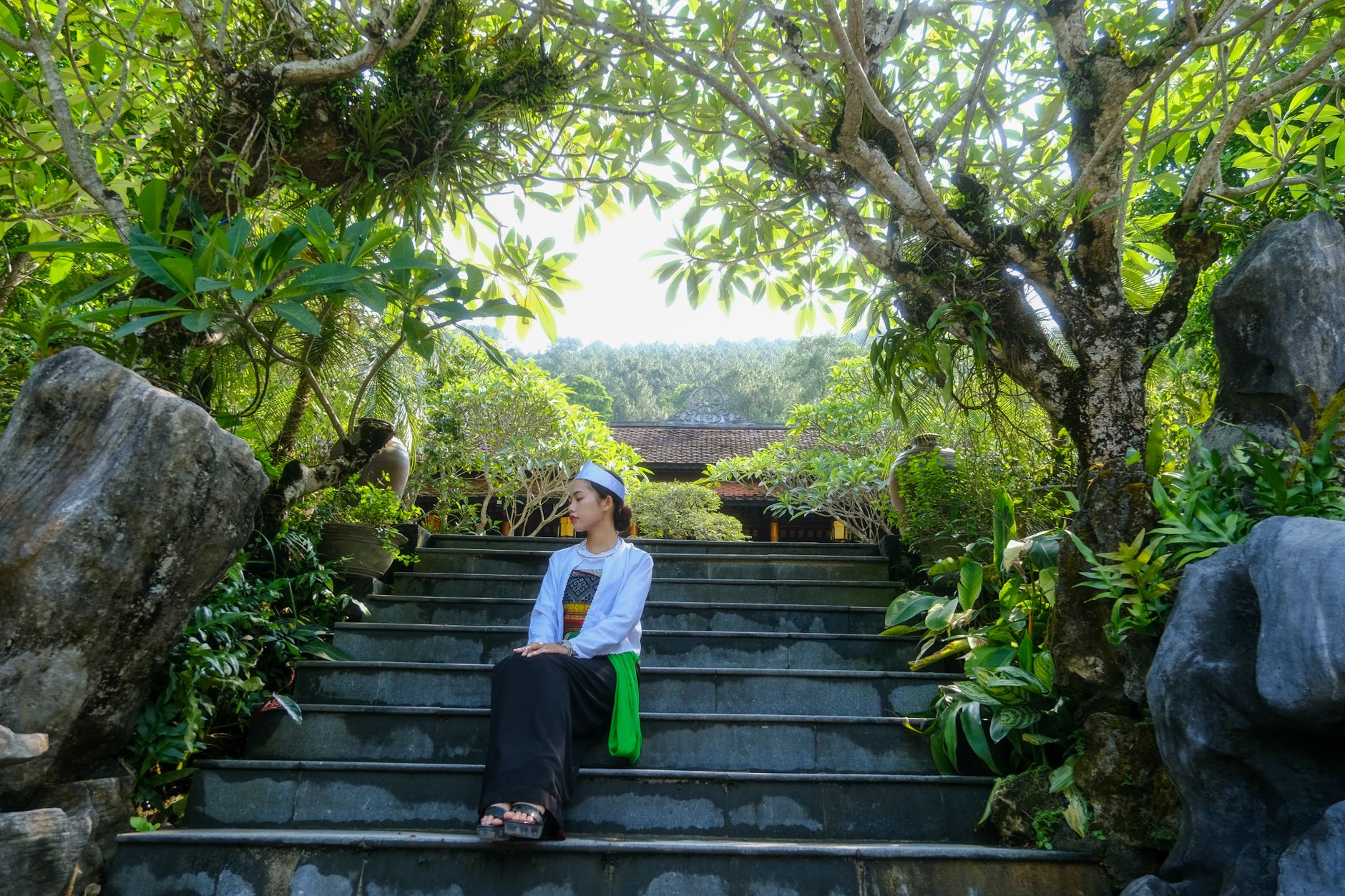 Woman in traditional attire sits peacefully on garden steps surrounded by lush greenery in Hue, Vietnam.