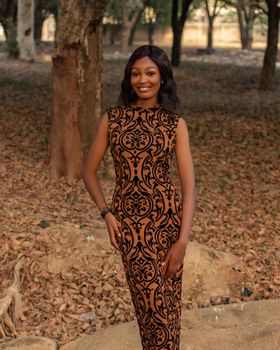 Stylish woman in a patterned dress posing confidently among autumn trees outdoors.
