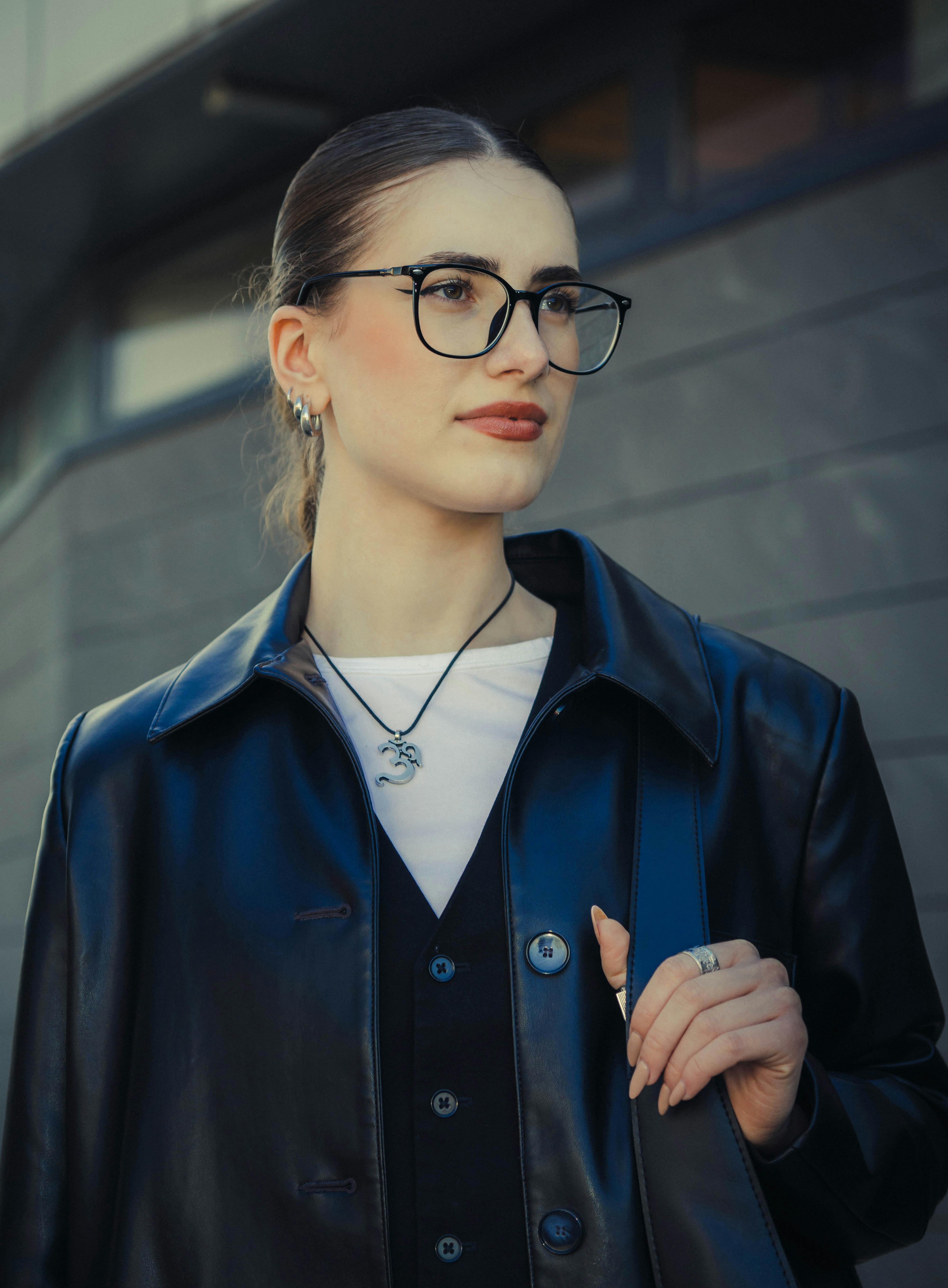 Безкоштовне Модна молода жінка в окулярах та шкіряній куртці біля сучасної будівлі. стокове фото