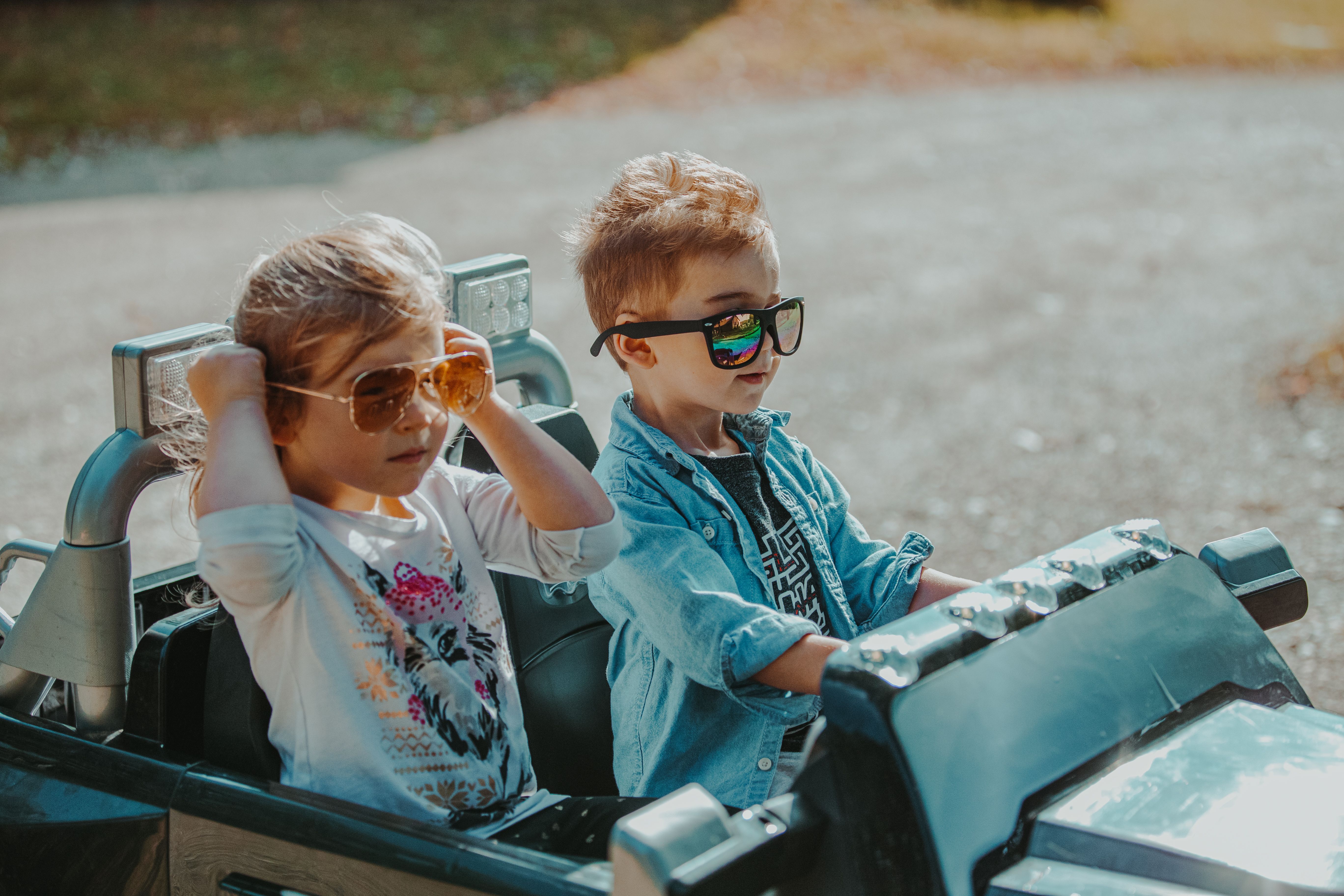 Two children wearing sunglasses enjoy driving a toy car outdoors in summer.