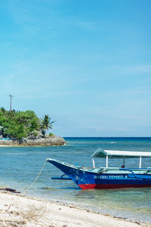 Free A serene view of a tropical beach with a traditional blue boat anchored near an island. Stock Photo