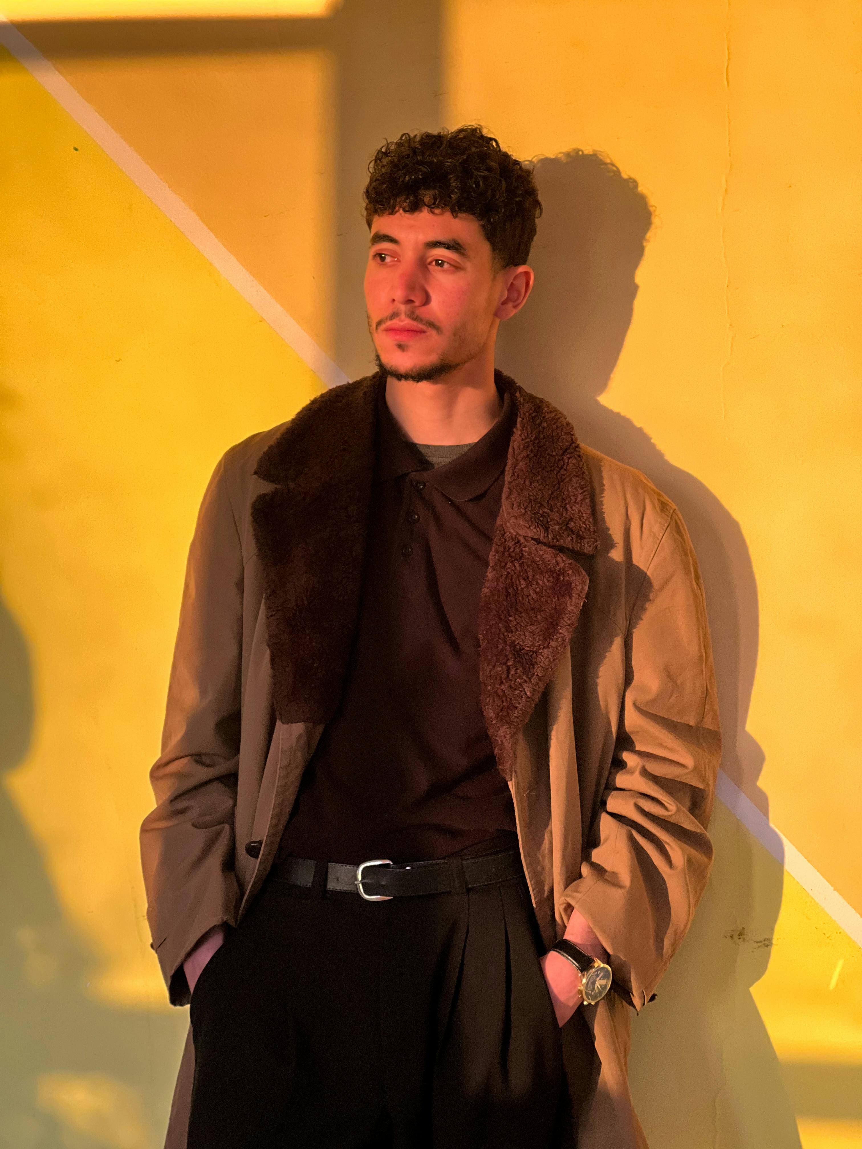 ingyenes Egy divatos fiatalember áll egy meleg, aranyfényű falnak támaszkodva, erős árnyékot vetve. Stockfotó