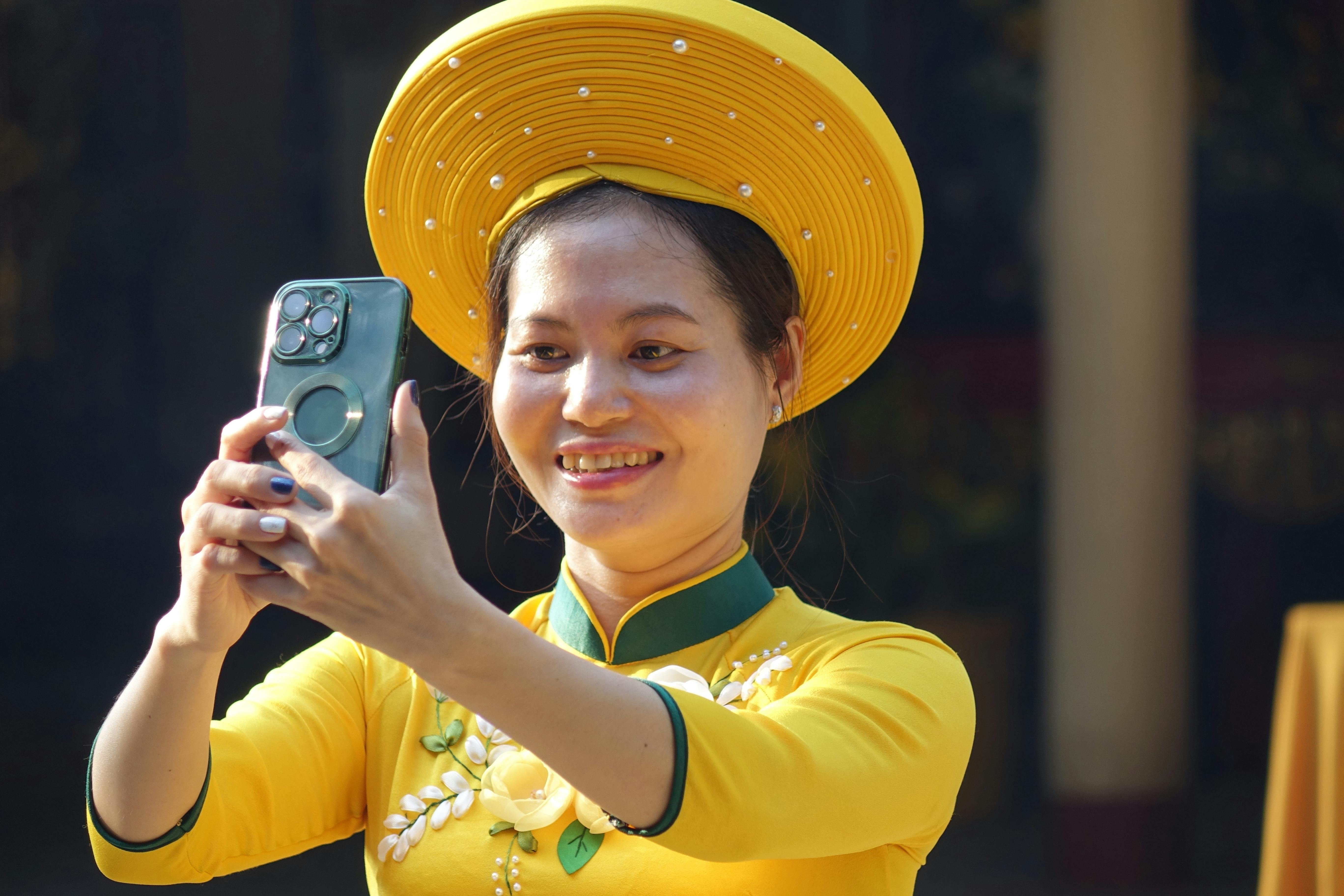 ฟรี หญิงสาวในชุดพื้นเมืองเวียดนามถ่ายเซลฟี่ด้วยสมาร์ทโฟนกลางแจ้ง คลังภาพถ่าย