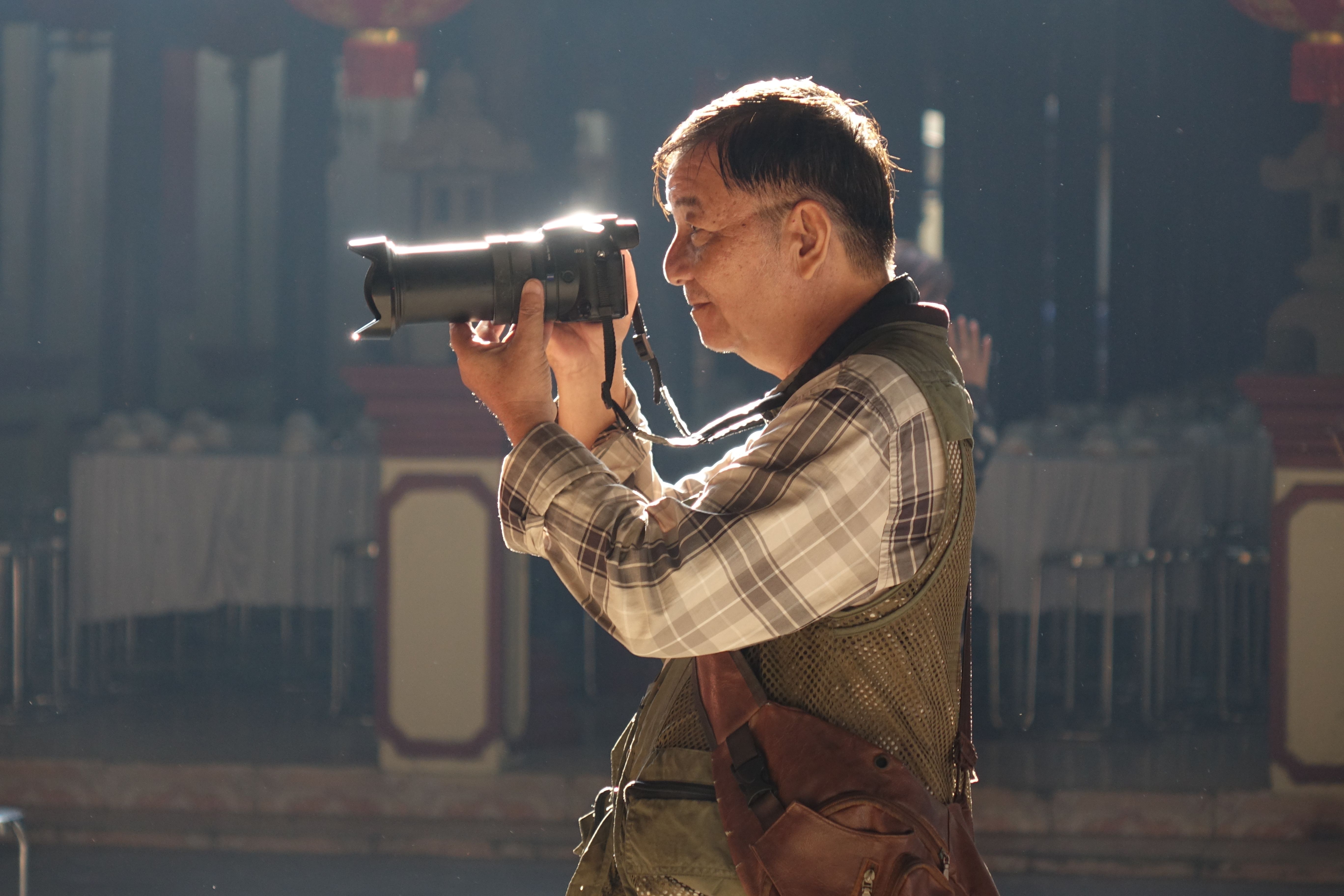 An elderly man taking photos indoors with a DSLR camera, highlighting passion for photography.