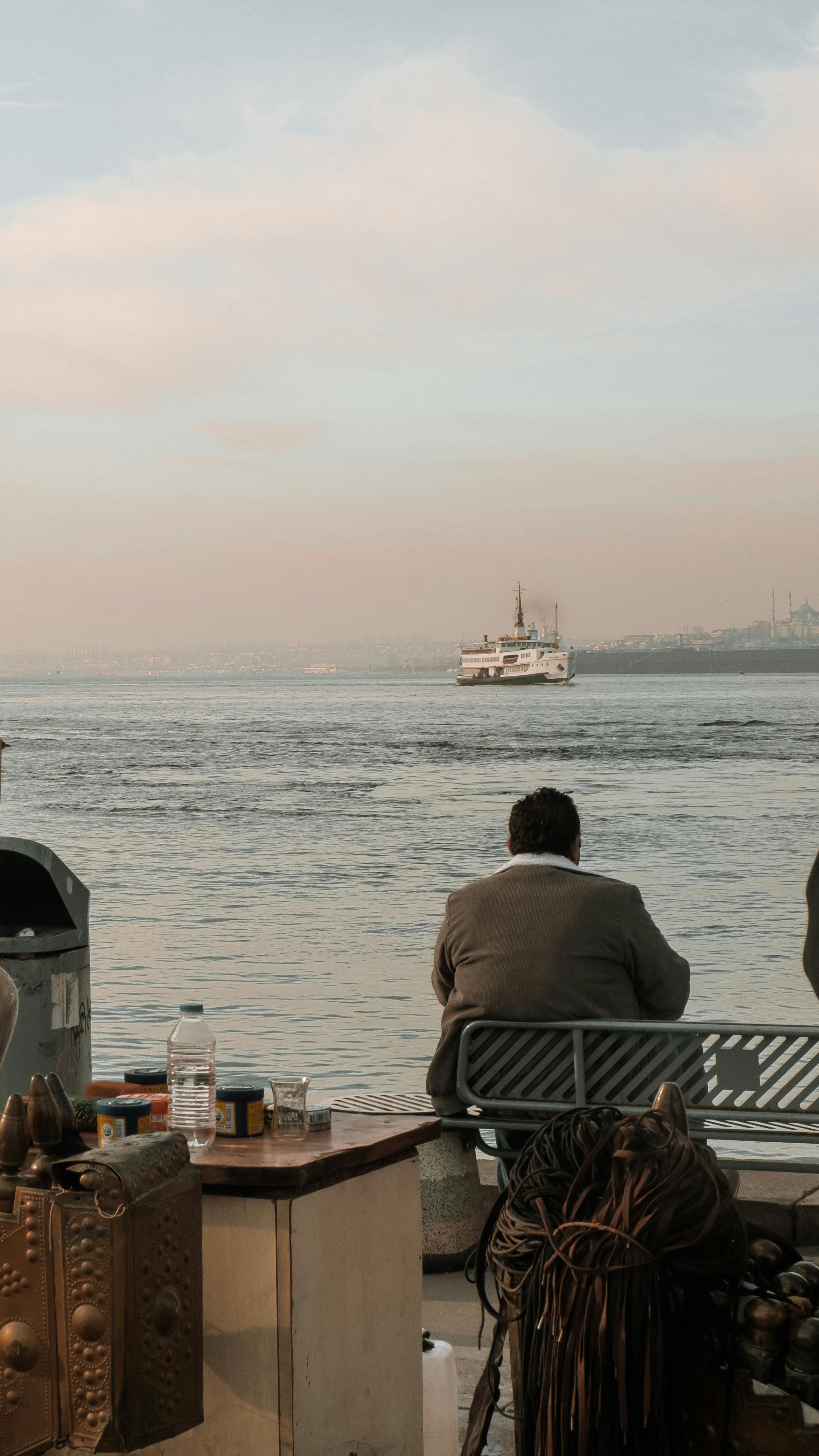 Безкоштовне Чоловік сидить біля річки Босфор, спостерігаючи за поромом, що пропливає повз, створюючи безтурботну атмосферу. стокове фото