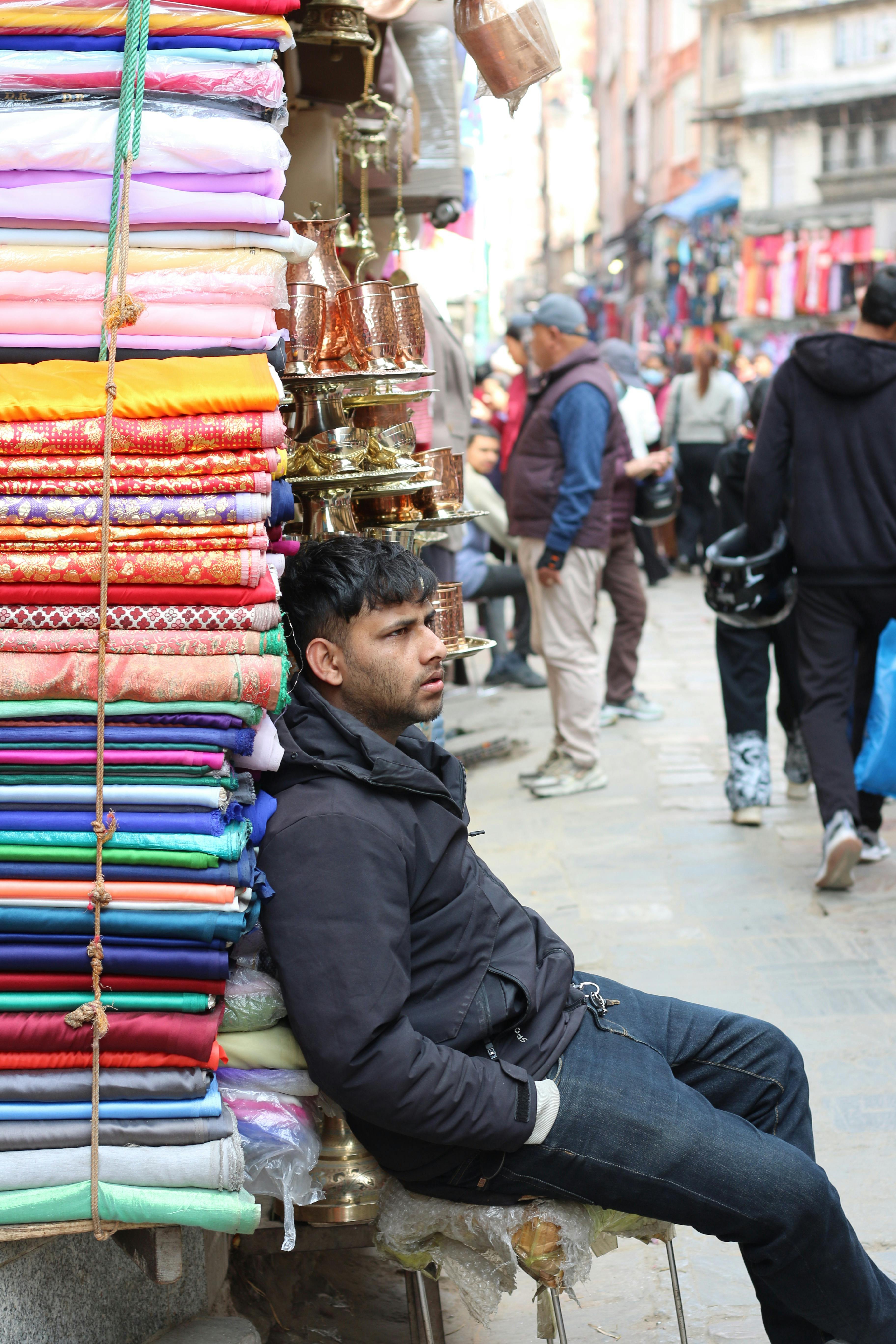免費 一名男子坐在加德滿都街頭，身旁擺放著色彩鮮豔的布料和黃銅製品，他用鏡頭捕捉熙熙攘攘的市場景象。 圖庫相片
