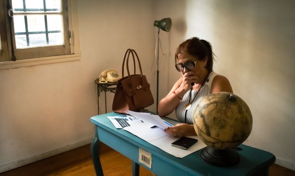 A woman focuses on paperwork at a desk with a globe and vintage phone.