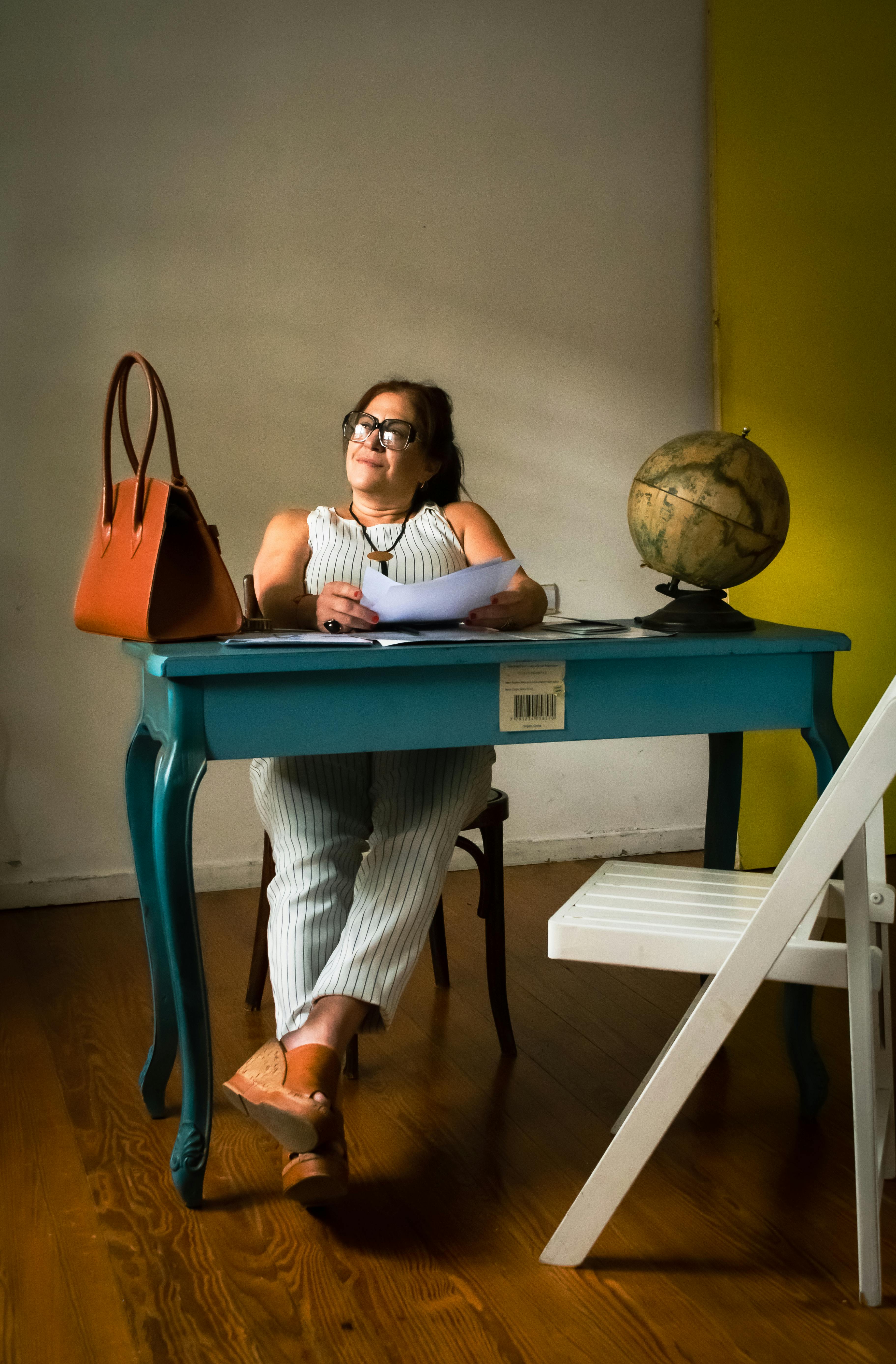 Безкоштовне Жінка сидить за синім столом з глобусом, заглиблена в думки у своєму домашньому кабінеті. стокове фото