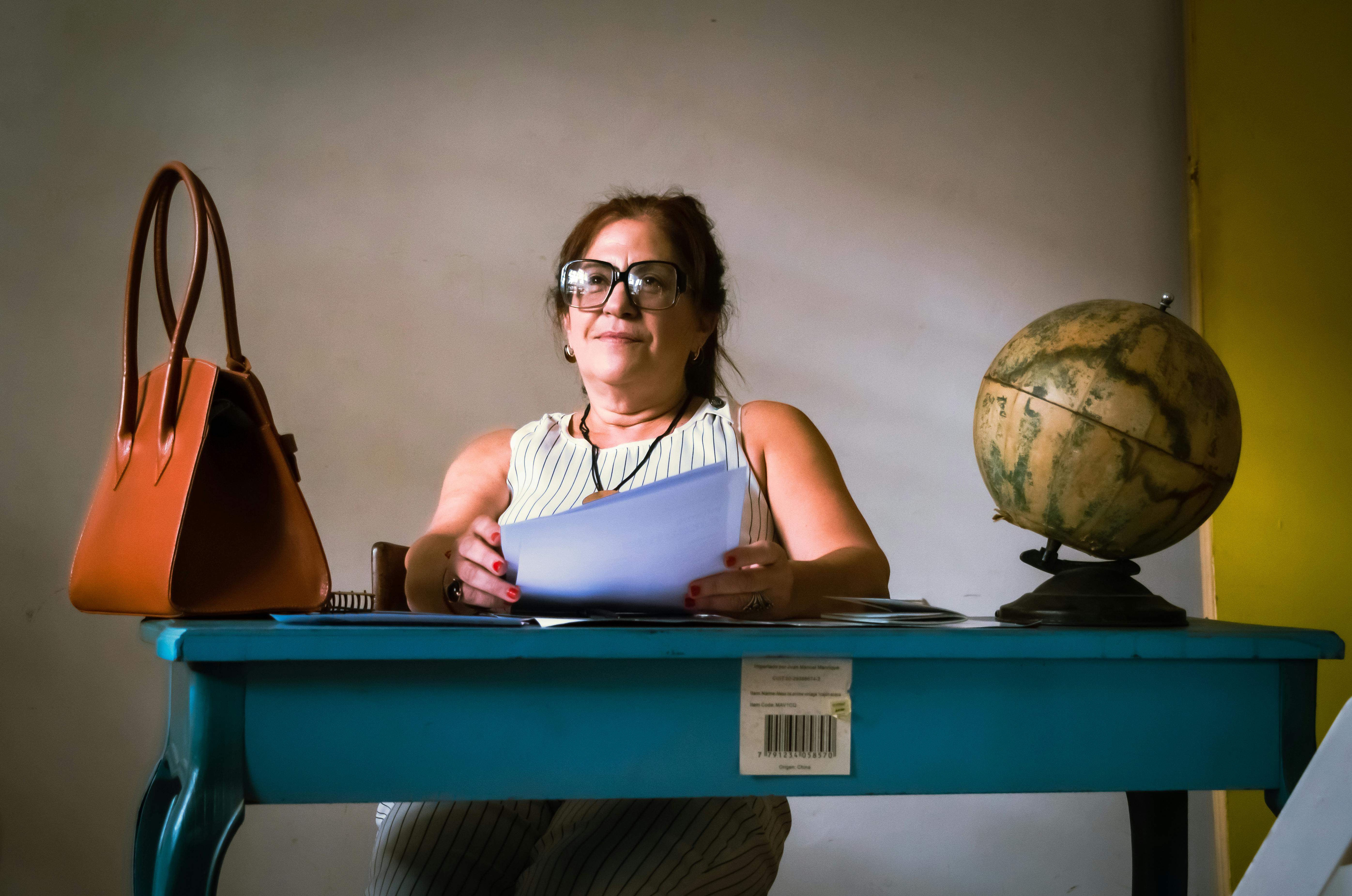 ฟรี หญิงสาวผู้มั่นใจนั่งอยู่ที่โต๊ะทำงานพร้อมเอกสาร ลูกโลก และกระเป๋าถือ สร้างบรรยากาศที่เป็นมืออาชีพภายในอาคาร คลังภาพถ่าย