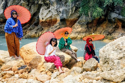 Four women with parasols sit on rocks by a river in West Java, Indonesia.