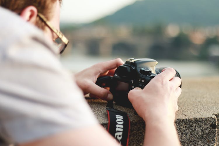 Man In White Top Holding Black Canon Dslr Camera On Gray Concrete Ledge