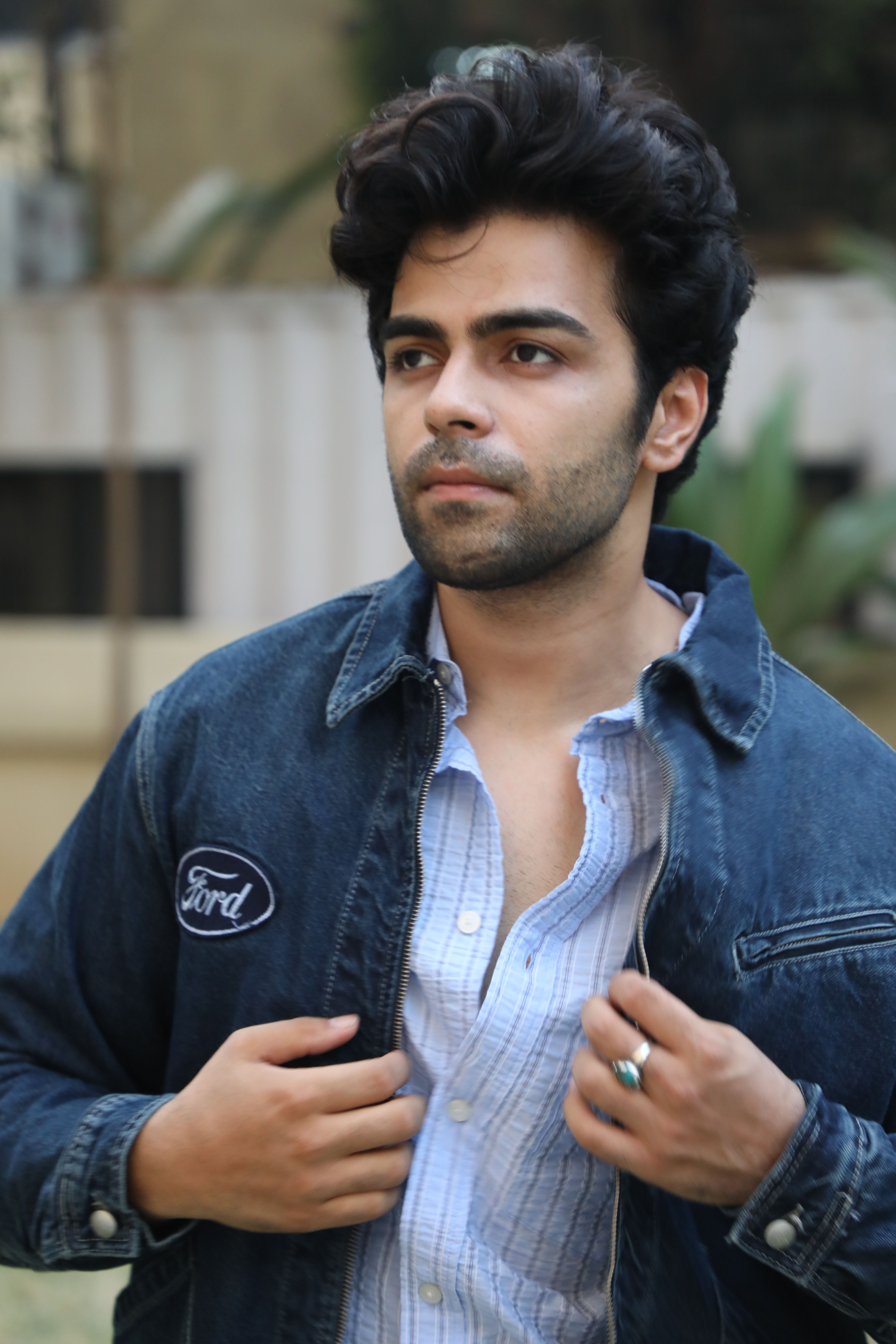 A young man posing confidently outdoors in a blue denim jacket and casual shirt.