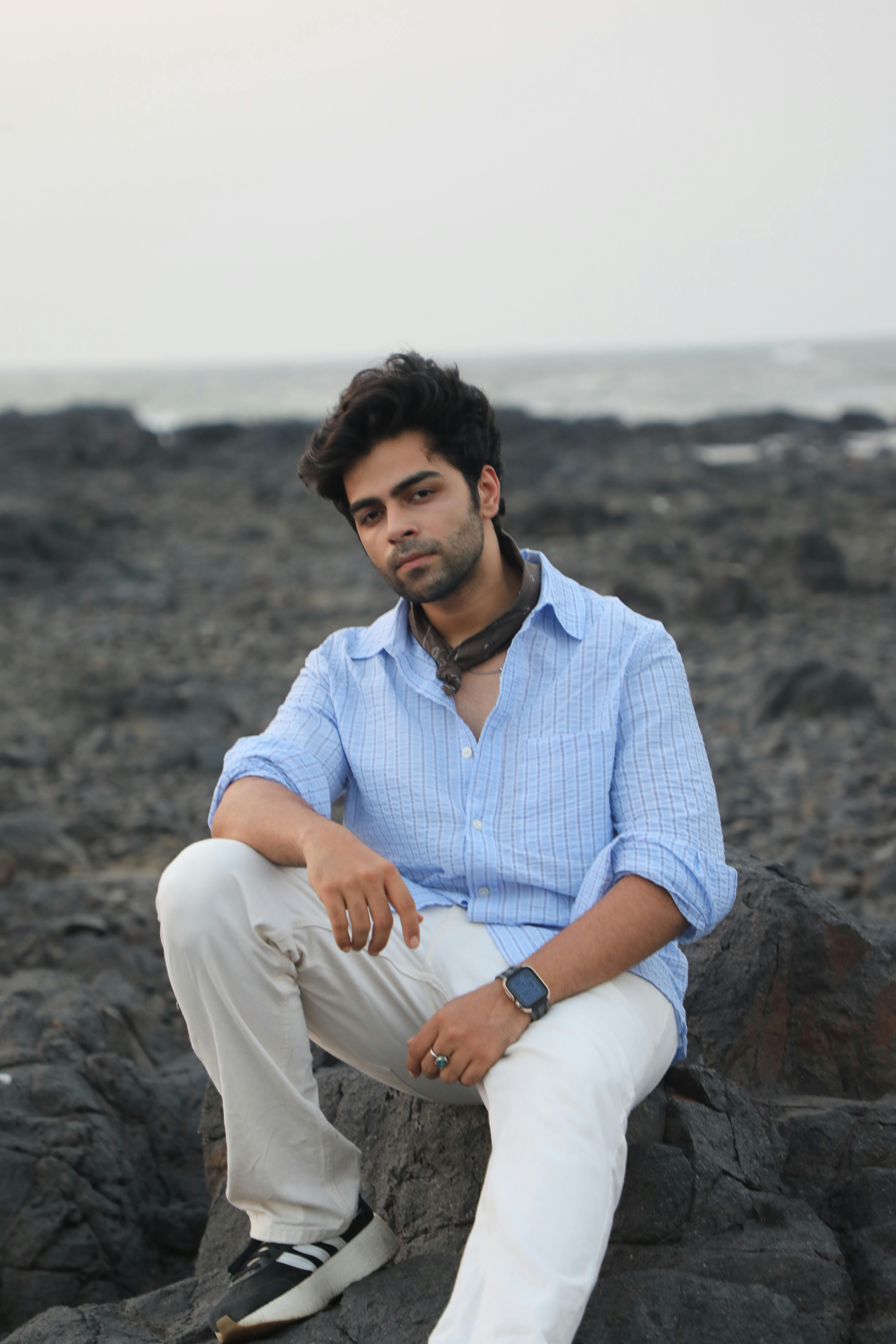 Young man in casual attire sits on a rocky beach during daytime, exuding a relaxed vibe.