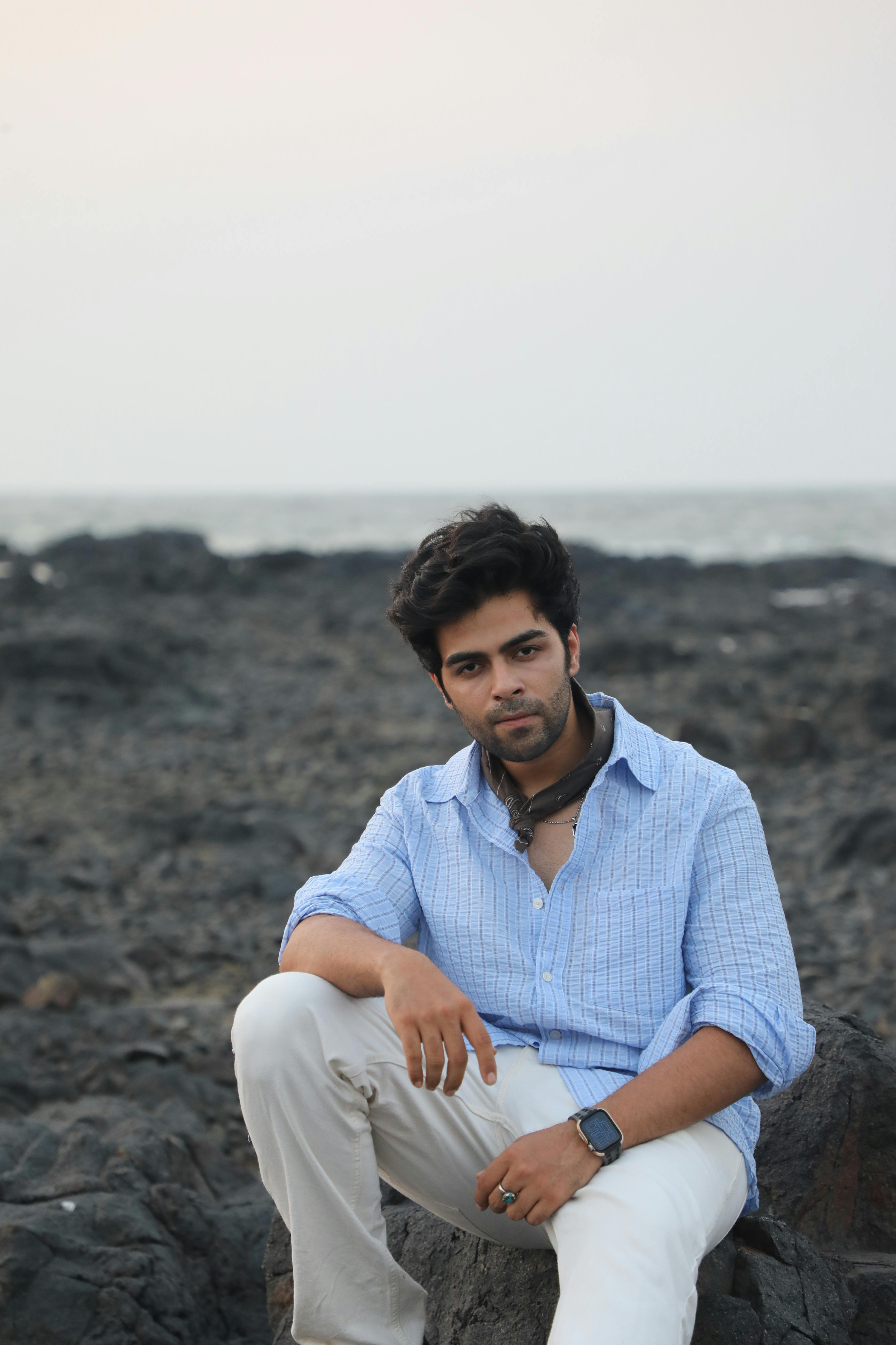 Young man in a casual blue shirt sitting on rocky beach at sunset.
