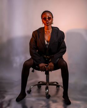 A stylishly dressed woman poses confidently seated on a chair in a studio.