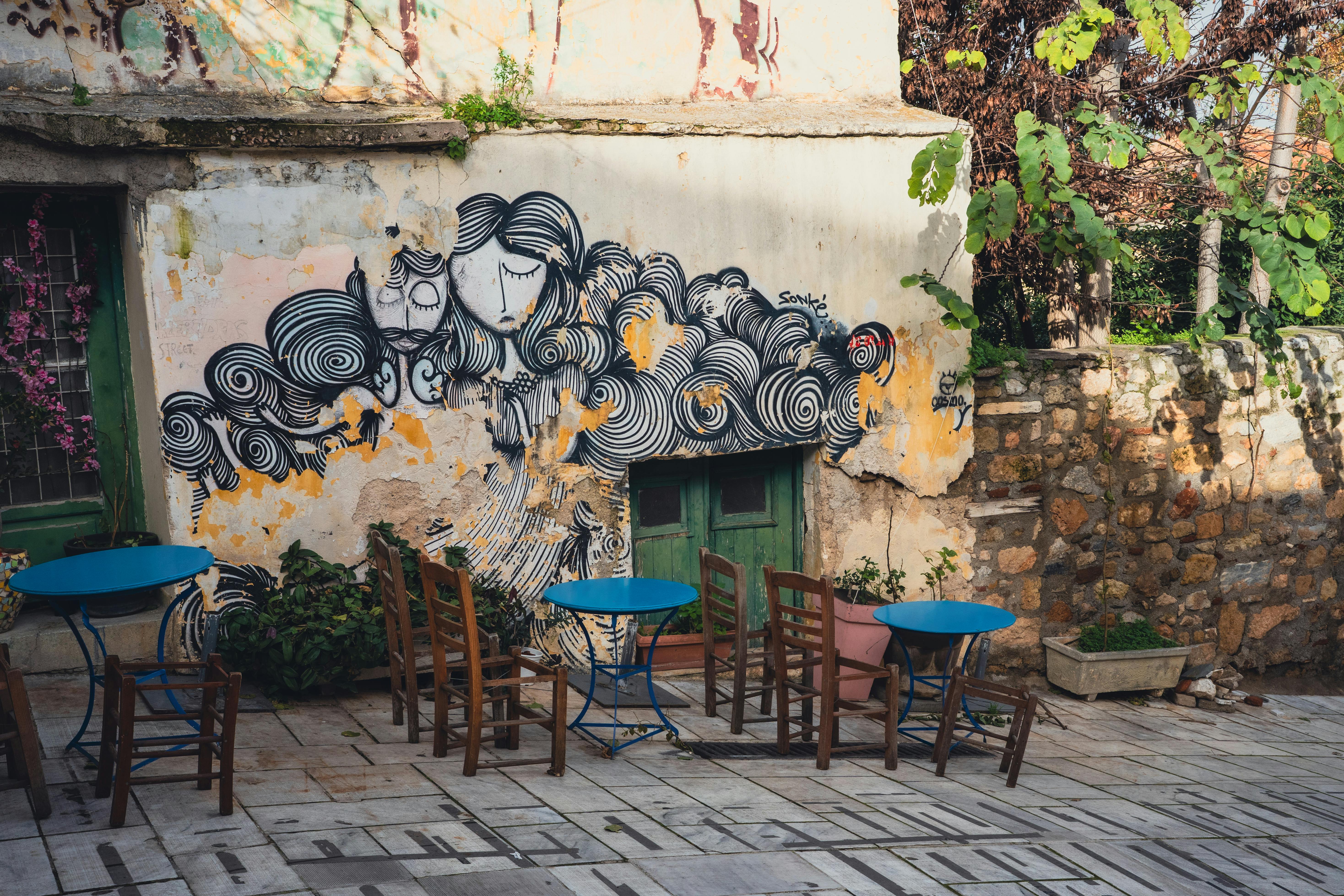 ingyenes Egy szabadtéri kávézó élénk színű kőfalfestményekkel, kék asztalokkal, üres székekkel és lombos növényekkel büszkélkedhet. Stockfotó
