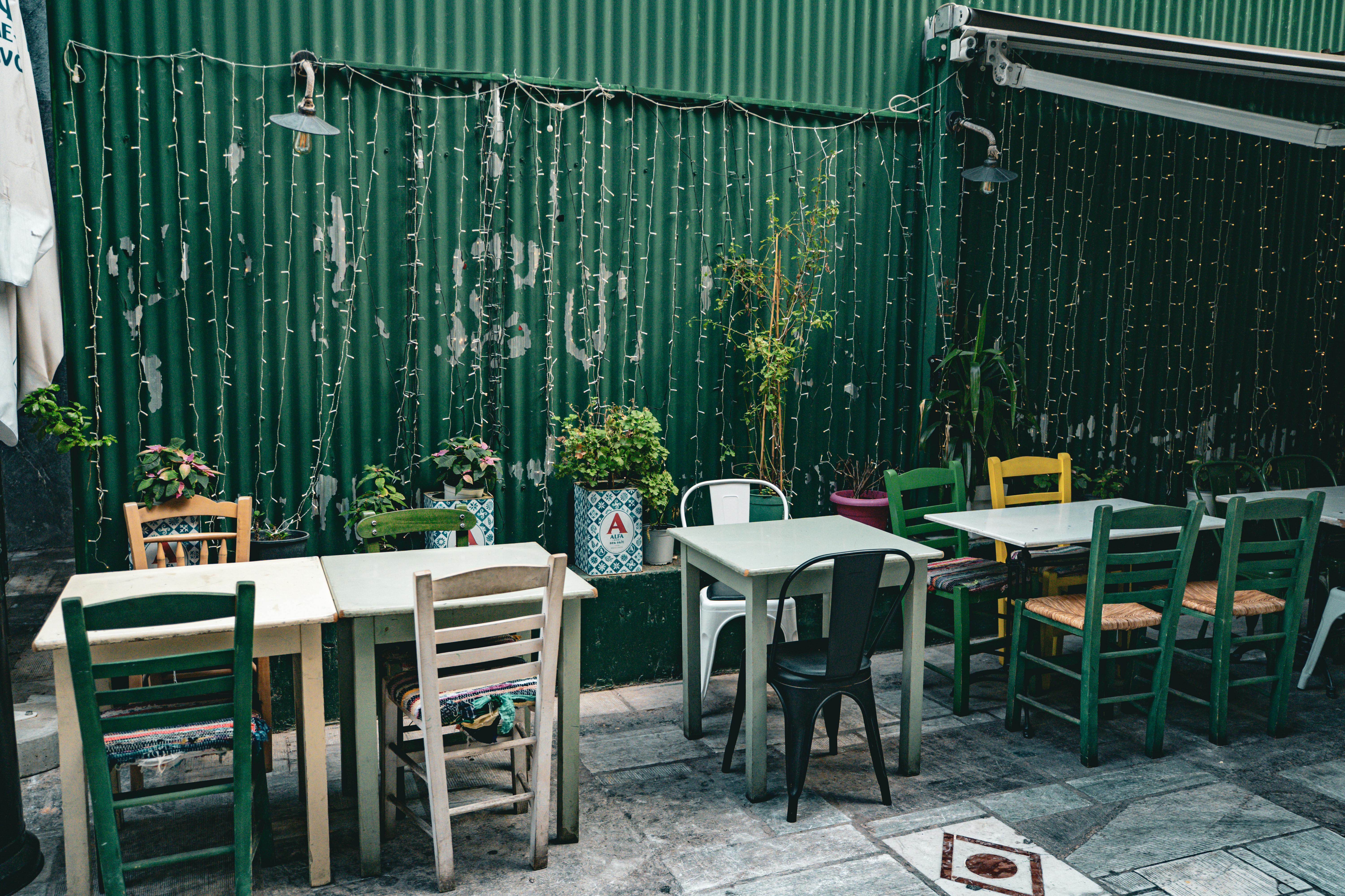 ฟรี คาเฟ่กลางแจ้งบรรยากาศอบอุ่น ตกแต่งด้วยเก้าอี้หลากหลายสไตล์และแสงไฟสลัวๆ บนฉากหลังสีเขียวแบบชนบท คลังภาพถ่าย