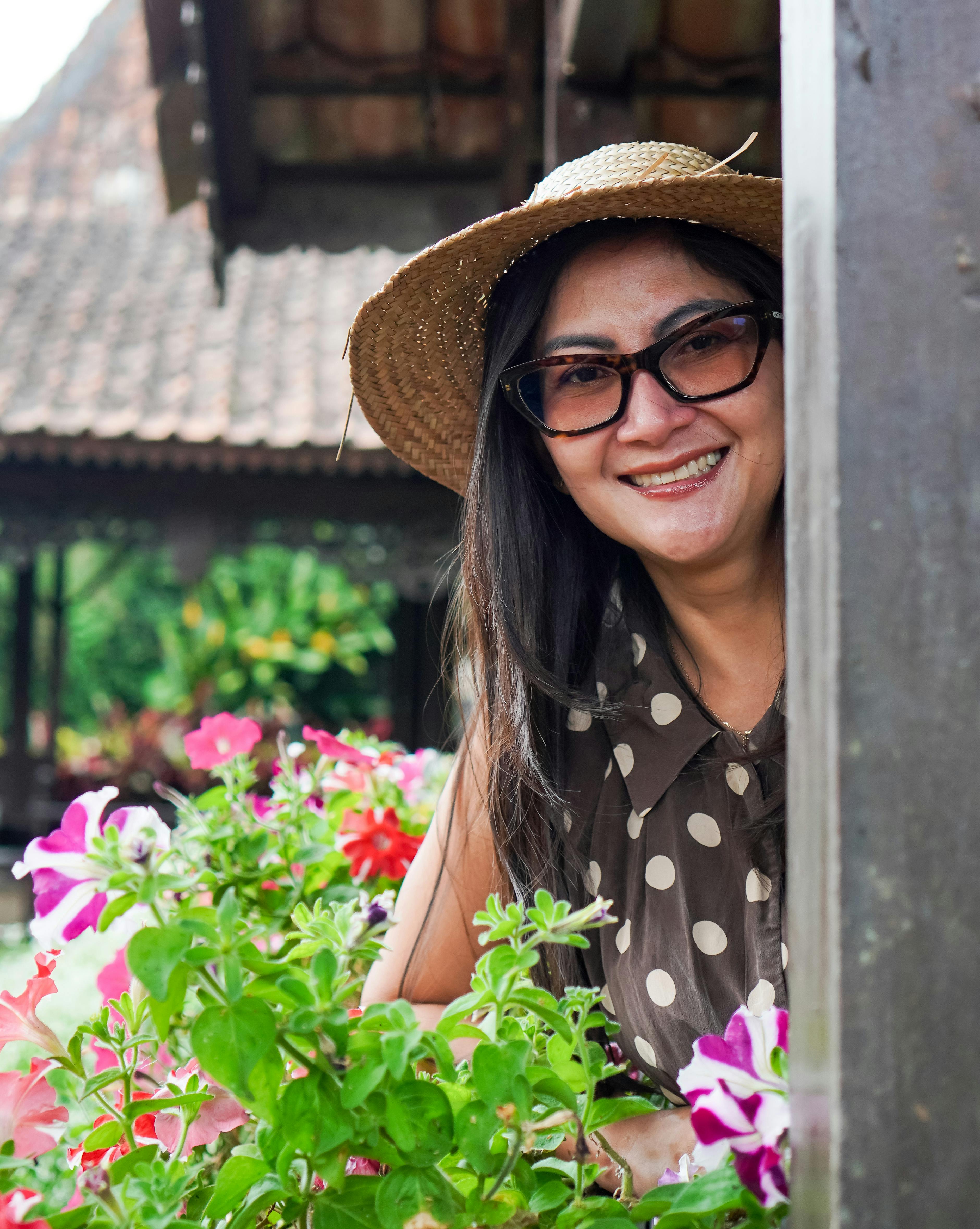 免費 一位頭戴草帽、笑容燦爛的女子在印尼的花園裡欣賞著五彩繽紛的花朵，展現出濃濃的夏日氣息。 圖庫相片