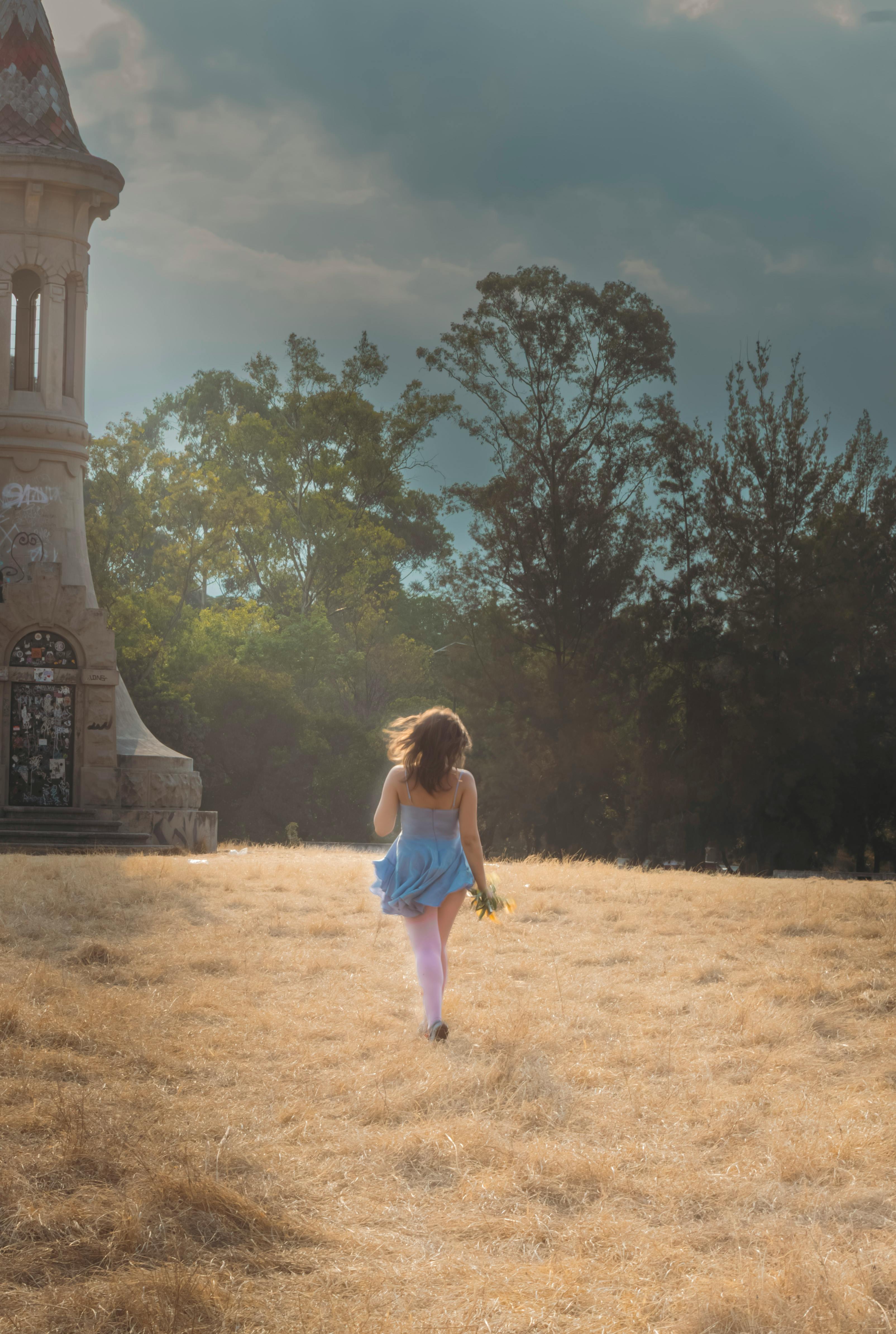 Безкоштовне Молода дівчина в синій сукні біжить сонячним полем, поруч з історичною вежею на тлі дерев. стокове фото