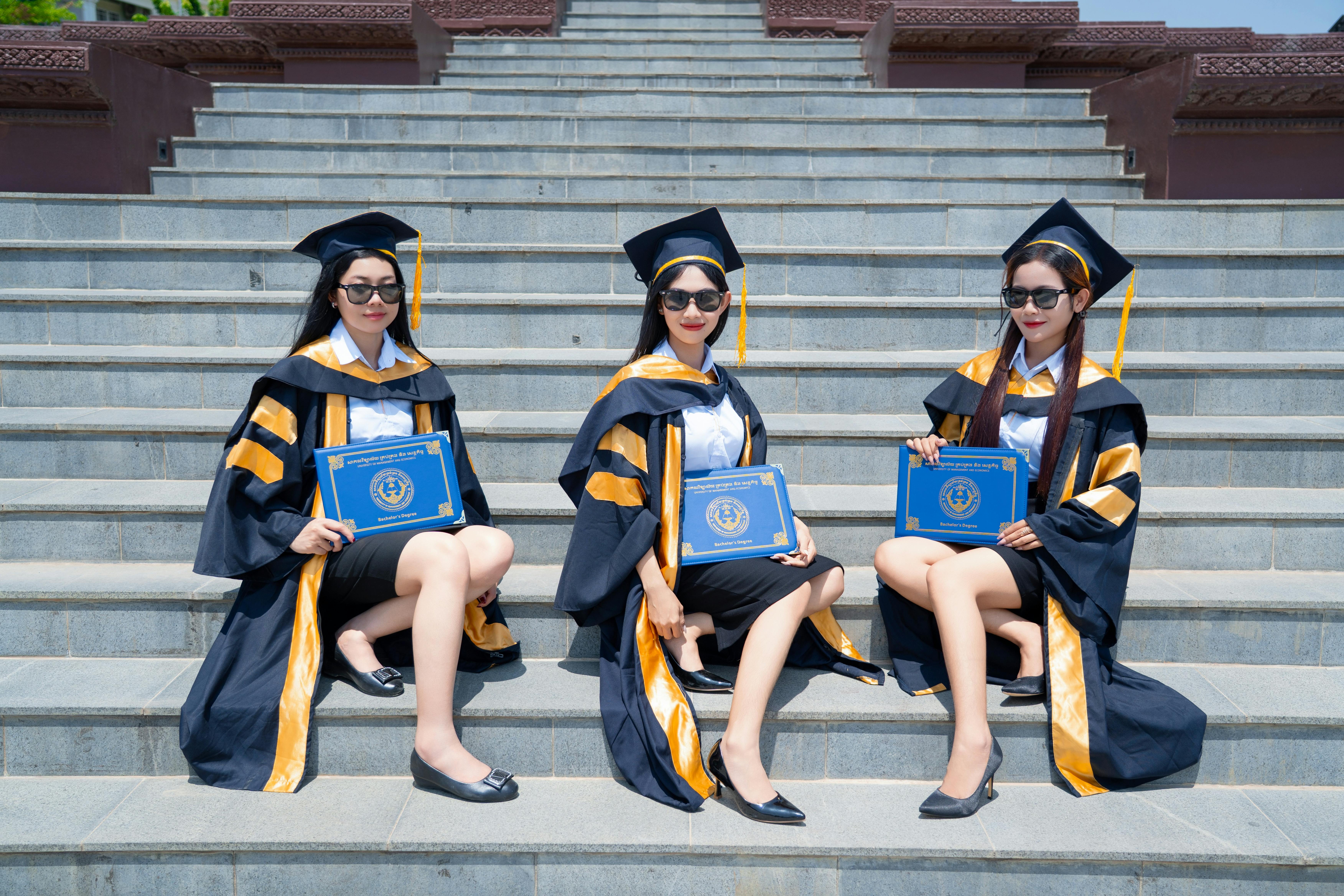 Безкоштовне Три випускниці у кепках та мантіях гордо демонструють свої дипломи на сходах кампусу. стокове фото