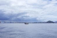 A tranquil view of a sailing ship and long bridge under a cloudy sky.