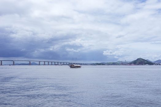 A tranquil view of a sailing ship and long bridge under a cloudy sky.