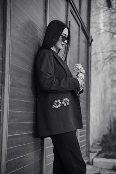 Sophisticated woman in sunglasses holding flowers, leaning against wooden wall.