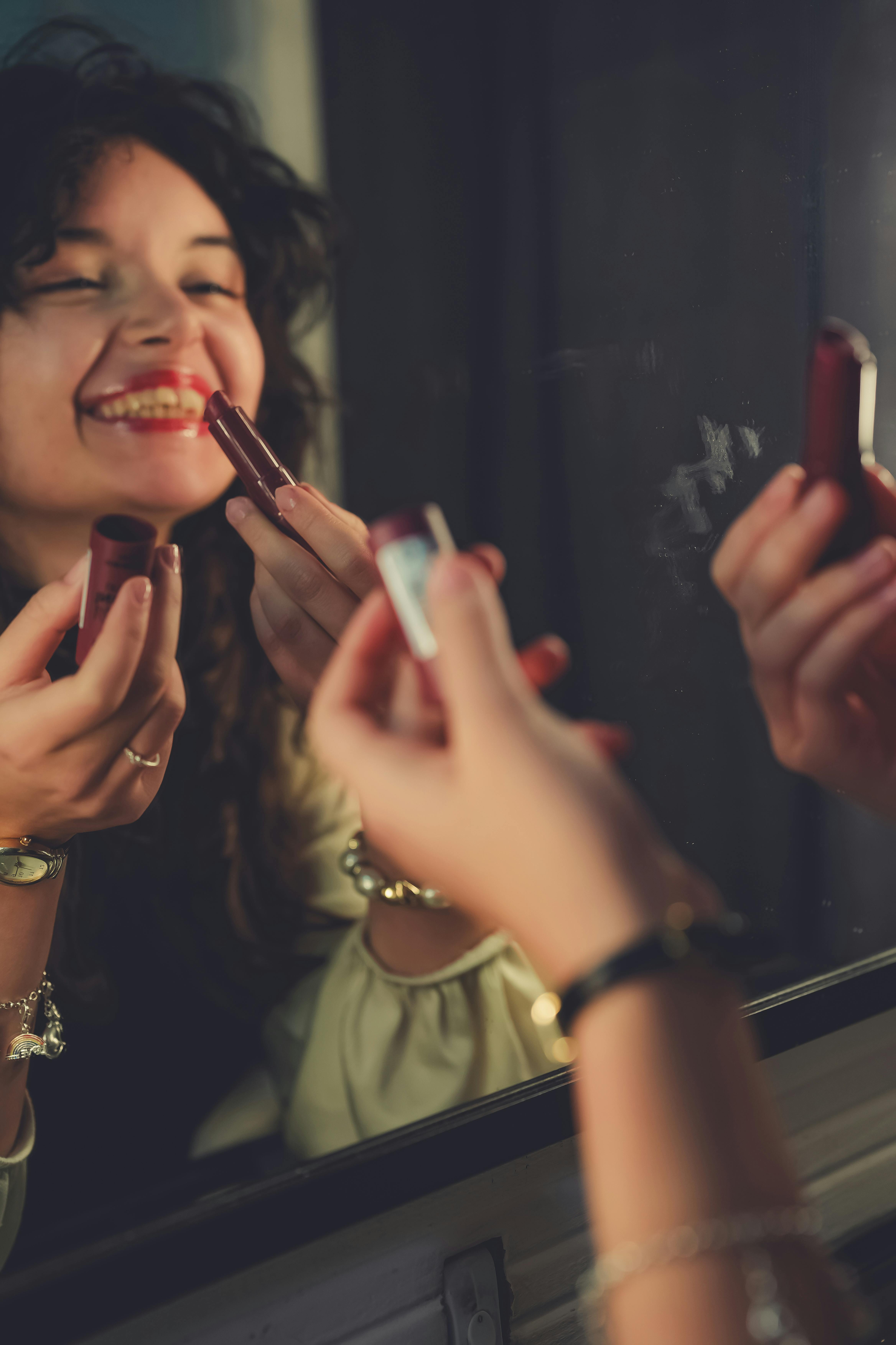 免費 一位微笑的女子在室內對著鏡子塗抹紅色唇膏。 圖庫相片