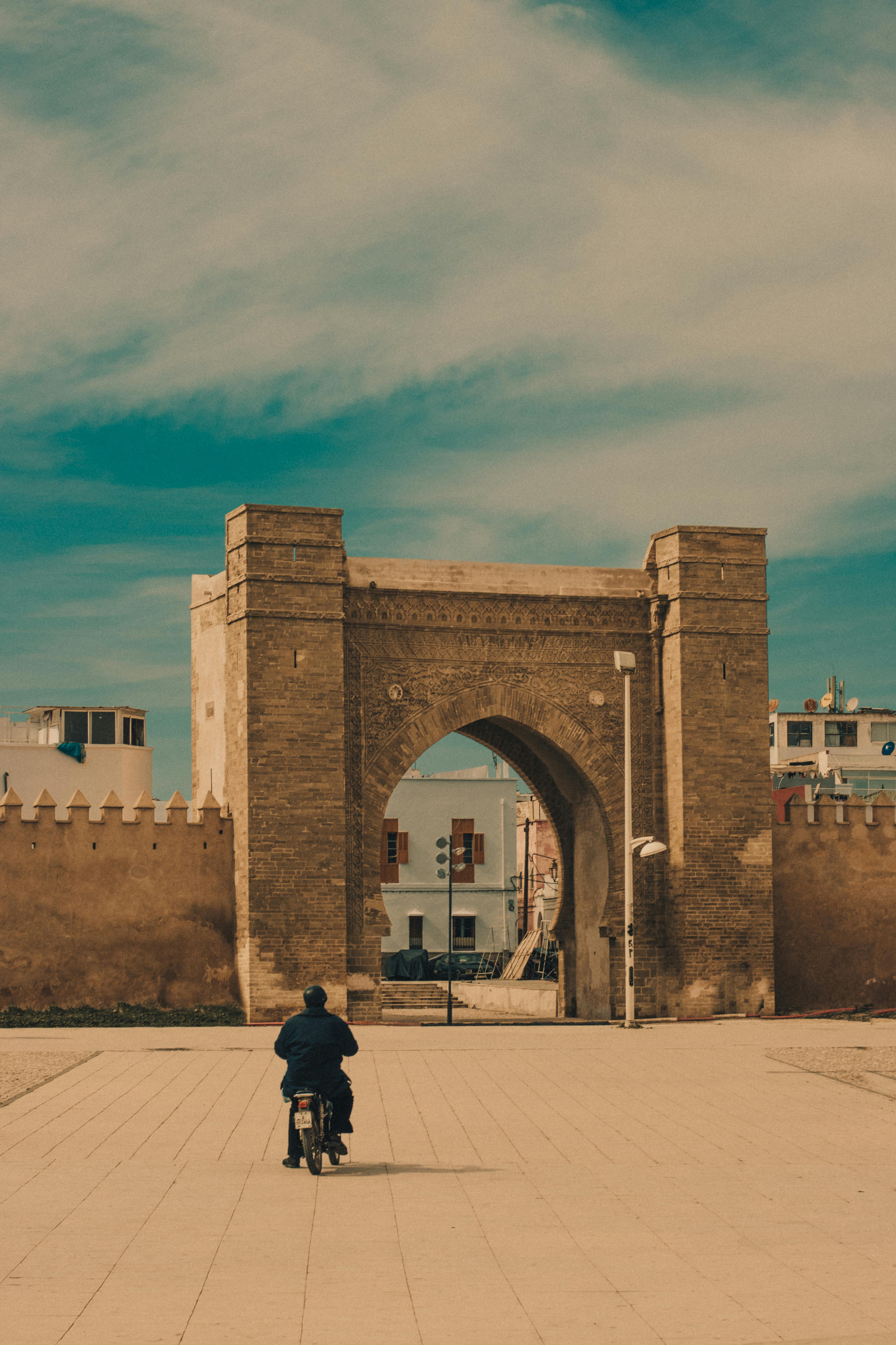 免費 藍天下，一名騎摩托車的男子獨自駛向一座歷史悠久的城門。 圖庫相片