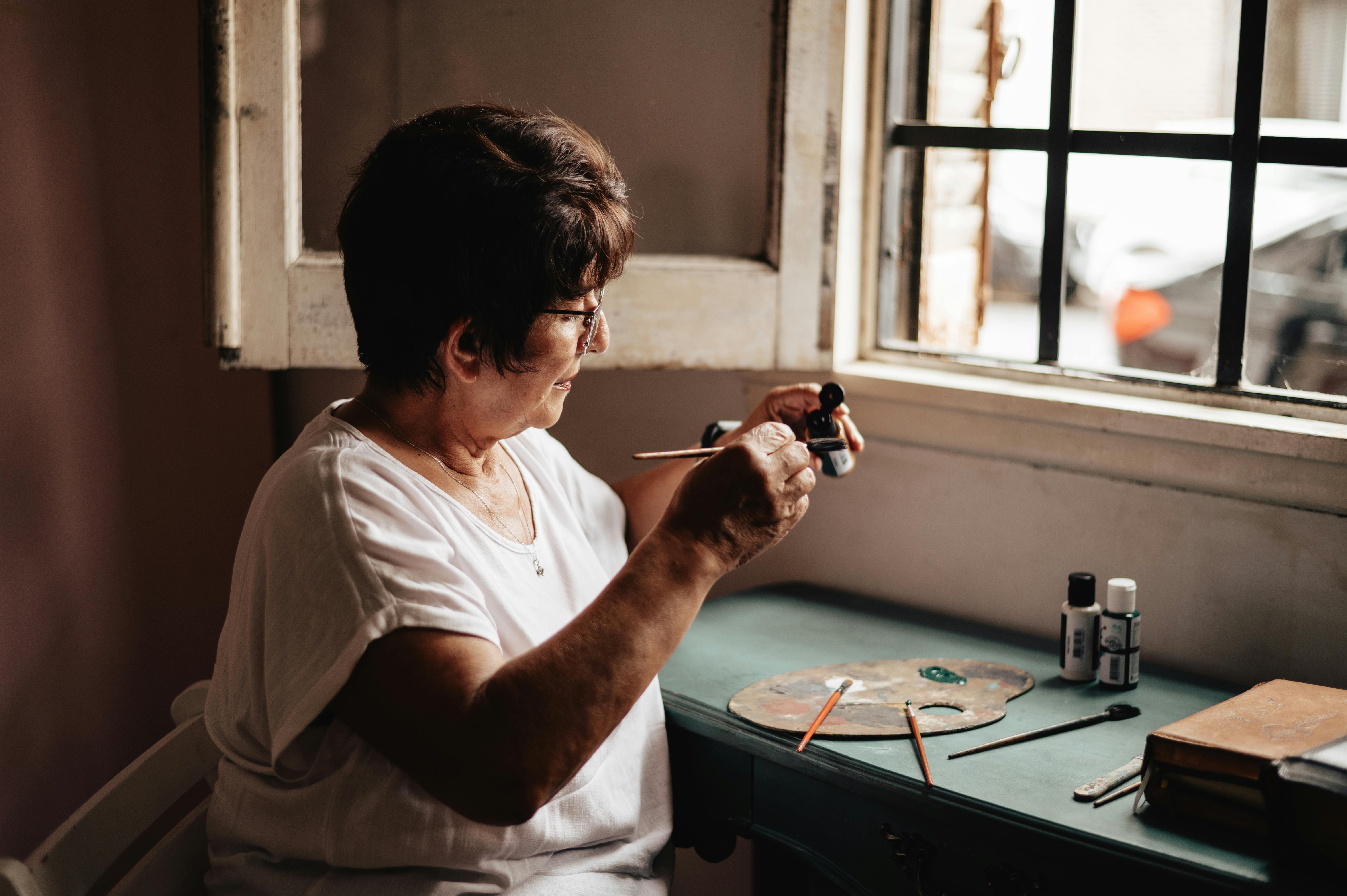 Безкоштовне Старша жінка займається малюванням біля вікна, з художнім приладдям на столі, створюючи мистецтво при природному світлі. стокове фото