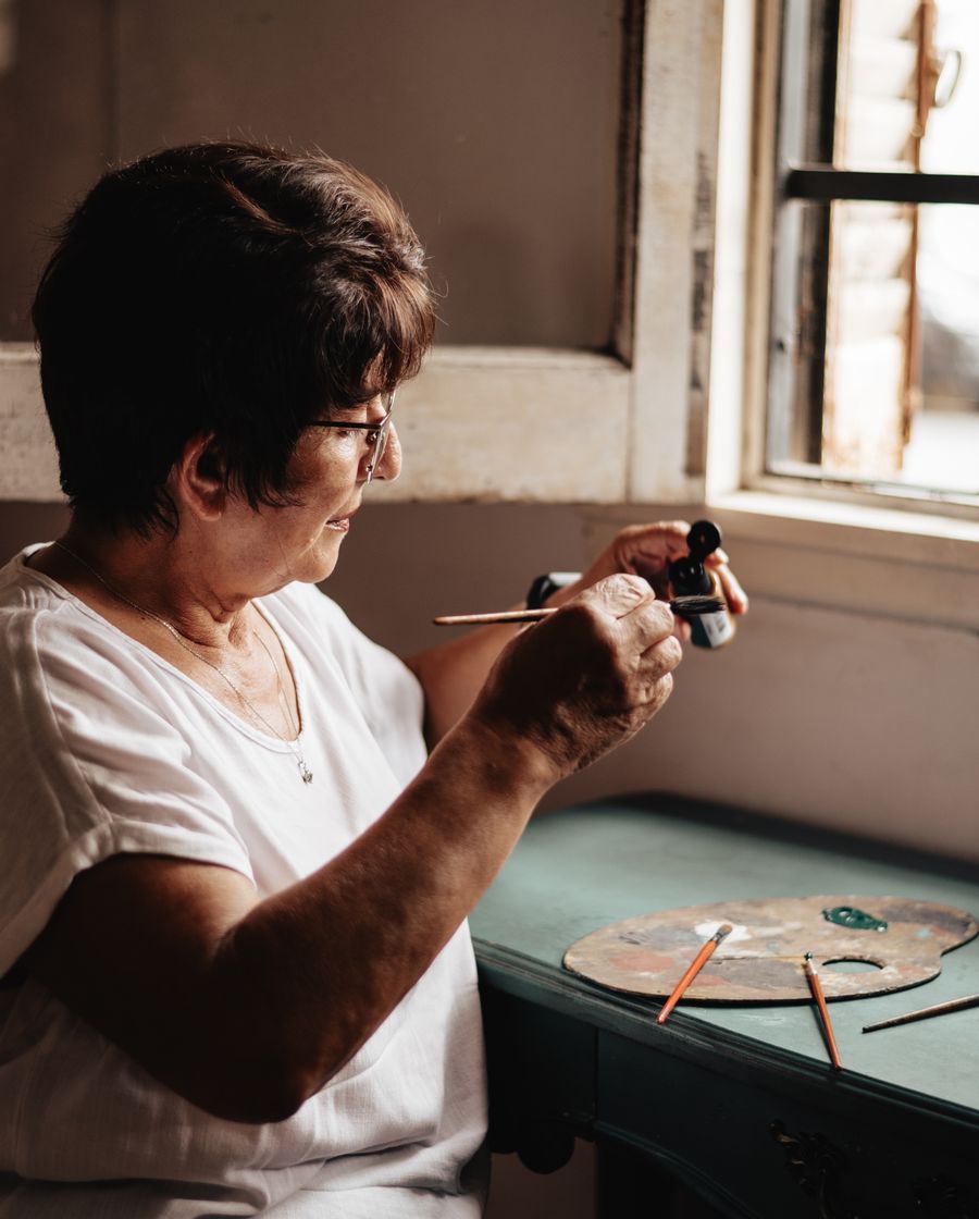 An older woman painting at a sunlit window, brushes and colours spread on the desk beside her.