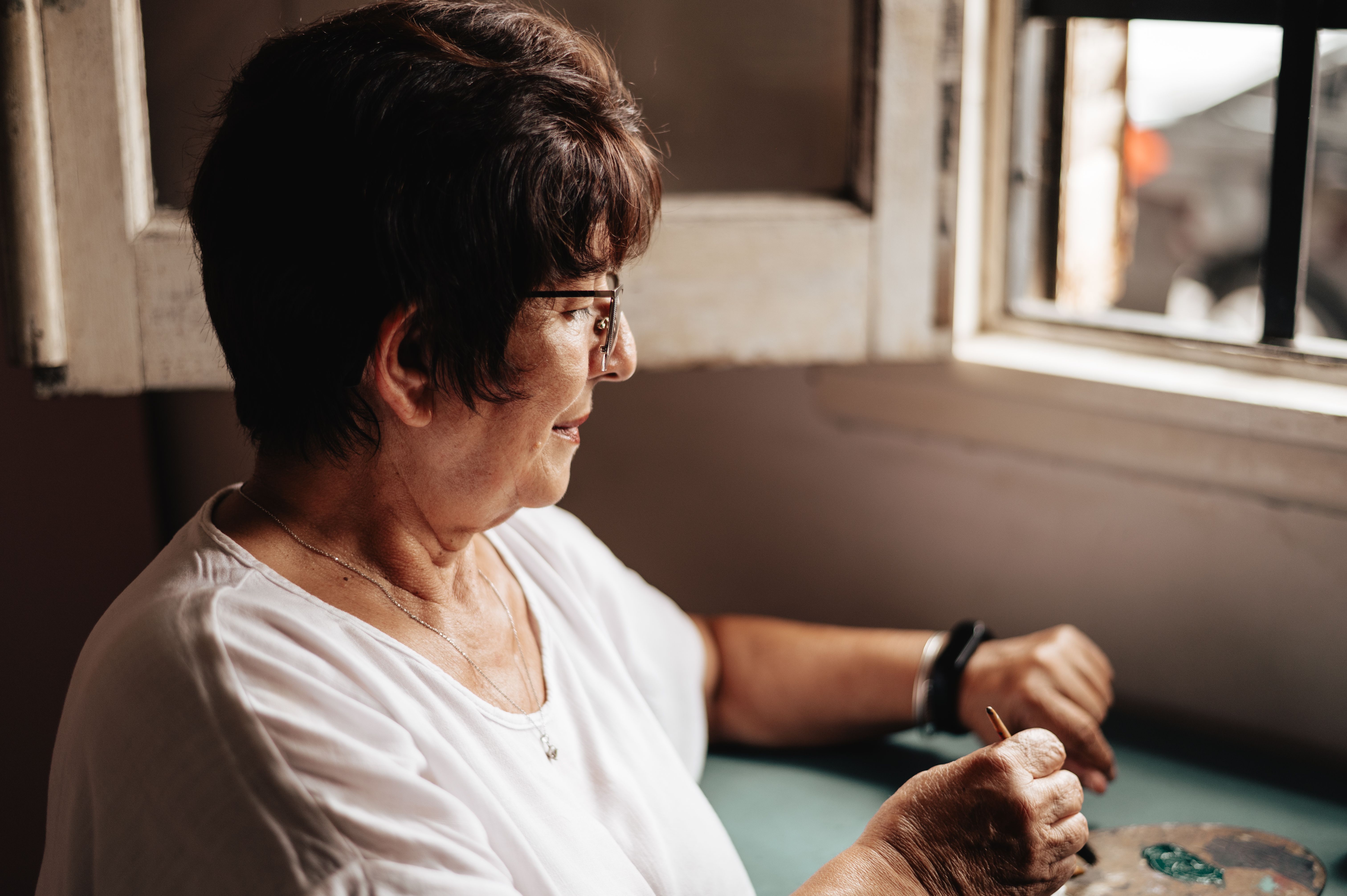 An elderly woman engaged in painting indoors by a window, bringing focus and creativity.