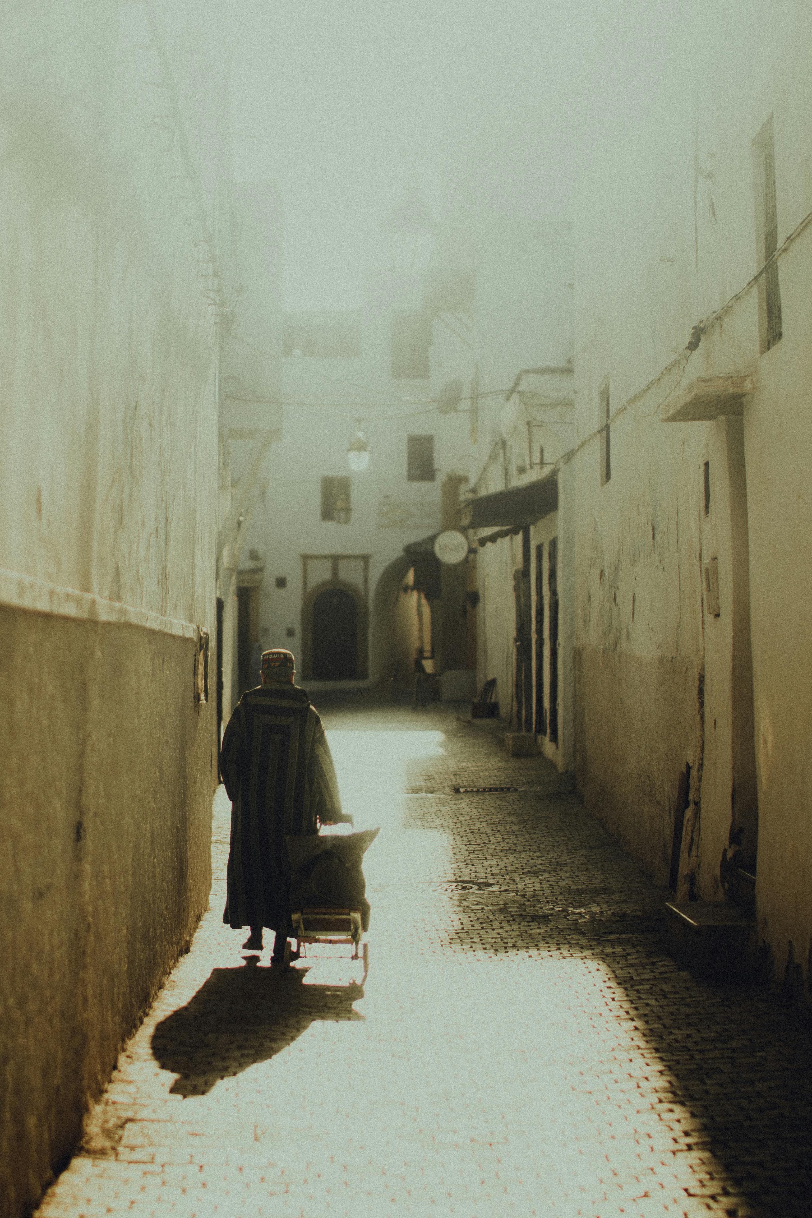 免費 一個孤獨的身影推著一輛小車走在陽光普照的小巷裡，投下長長的影子。 圖庫相片