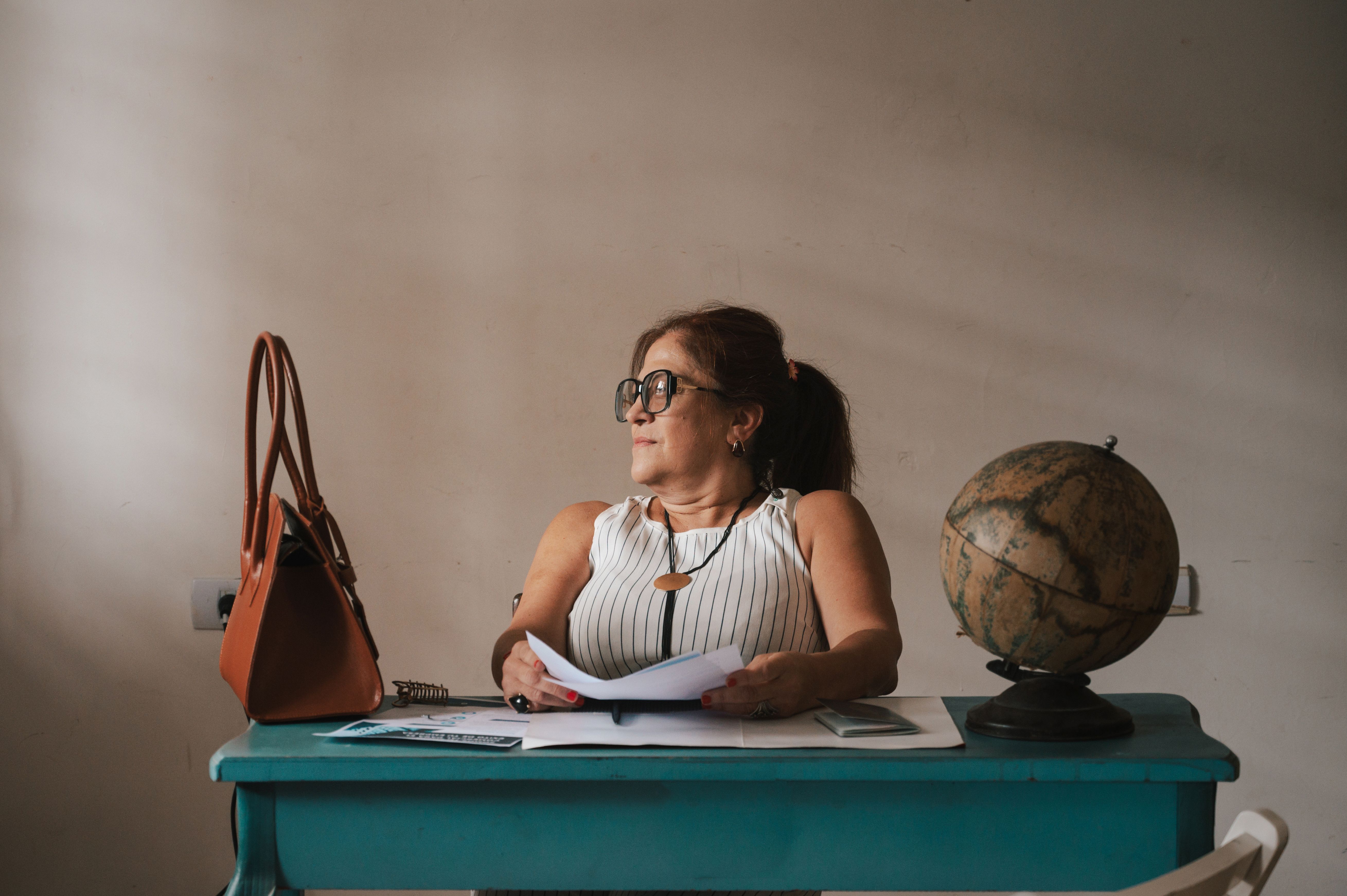 A woman sits at a vintage desk with a globe, documents, and a bag, evoking a scholarly atmosphere.