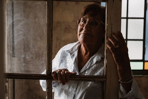 Free Portrait of an elderly woman looking through a rustic window in a vintage setting, evoking nostalgia. Stock Photo