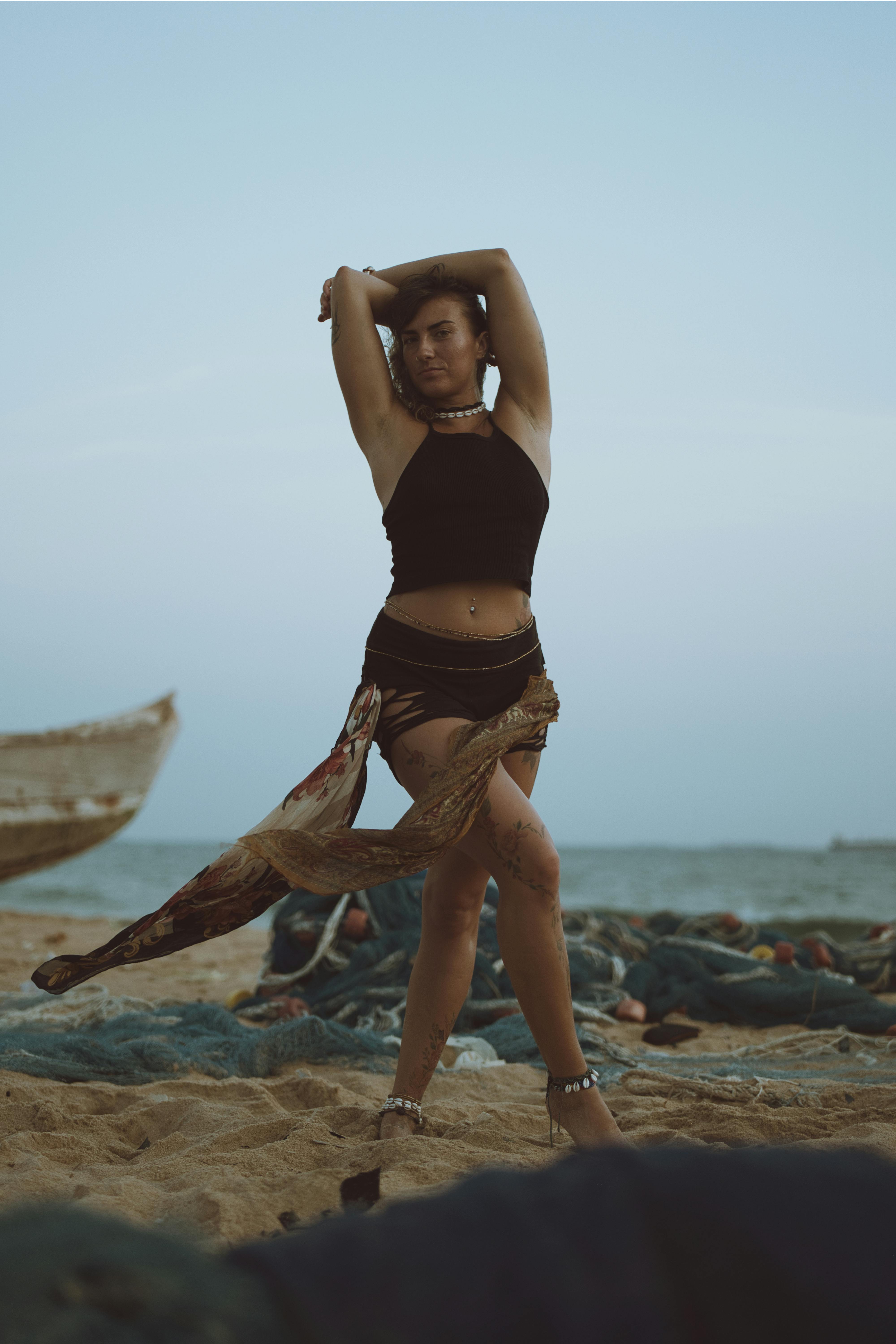 Безкоштовне Жінка в чорному топі та шортах впевнено позує на пляжі з мальовничим океанським фоном. стокове фото