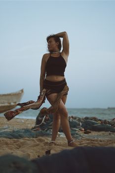 Fashionable woman striking a pose on a sandy beach in the evening, with scattered fishing nets and a boat in the background.