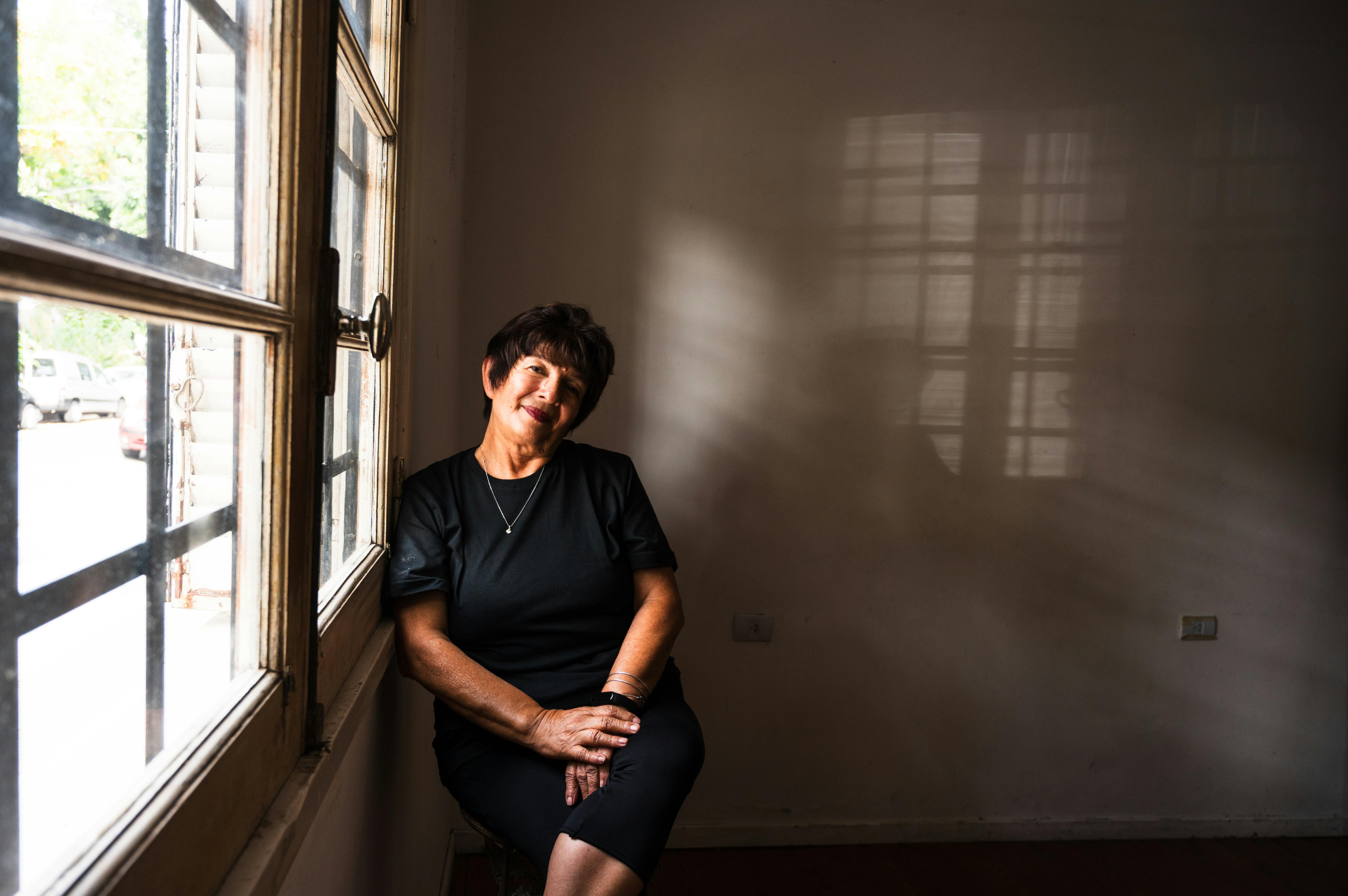 ฟรี ภาพถ่ายหญิงชรานั่งอยู่ข้างหน้าต่าง เงาอ่อนๆ ทอดลงบนผนัง คลังภาพถ่าย